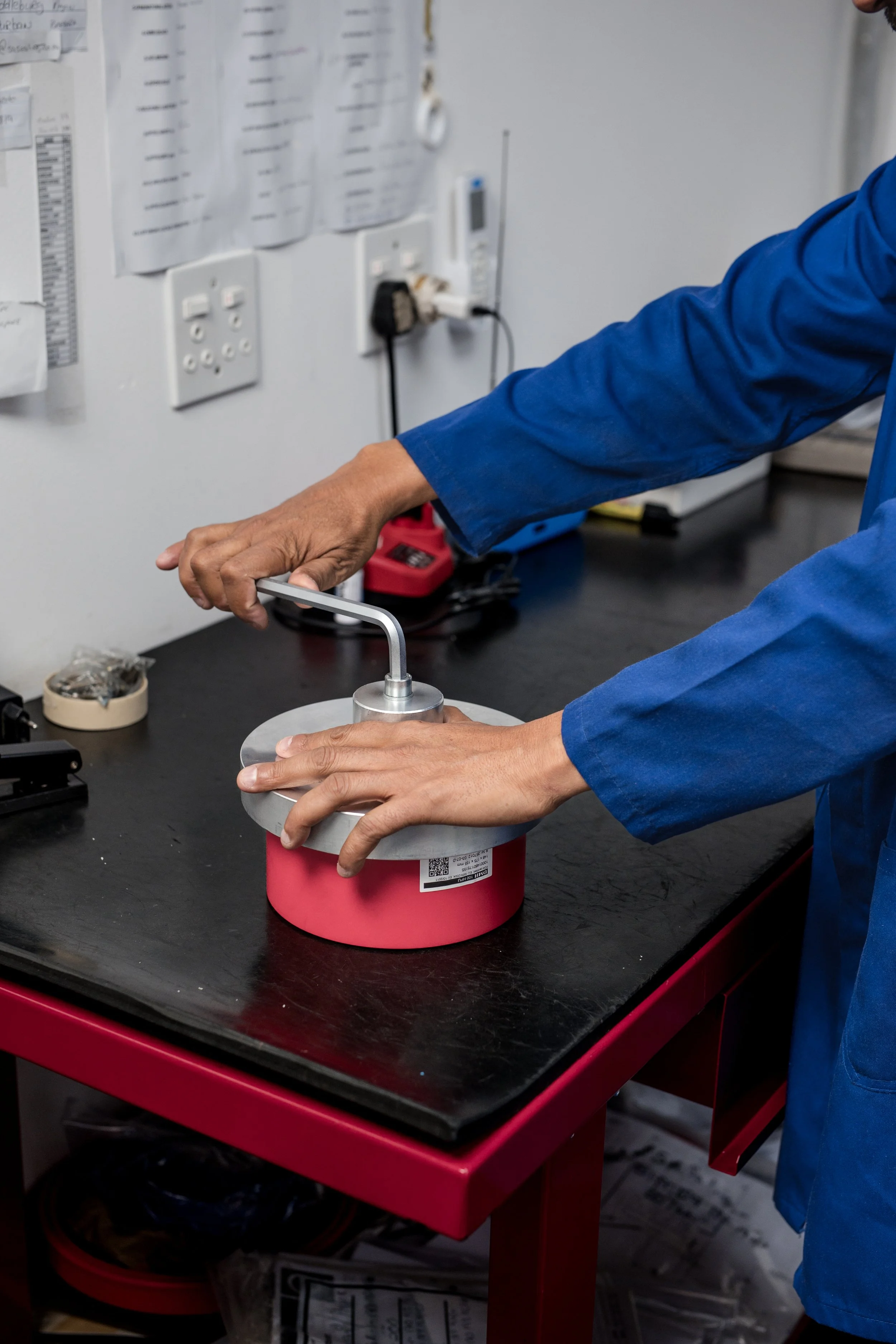 A person in a blue jacket working in a laboratory, handling a metal tool over a red and white container on a black workbench, with papers and laboratory equipment on the wall behind.