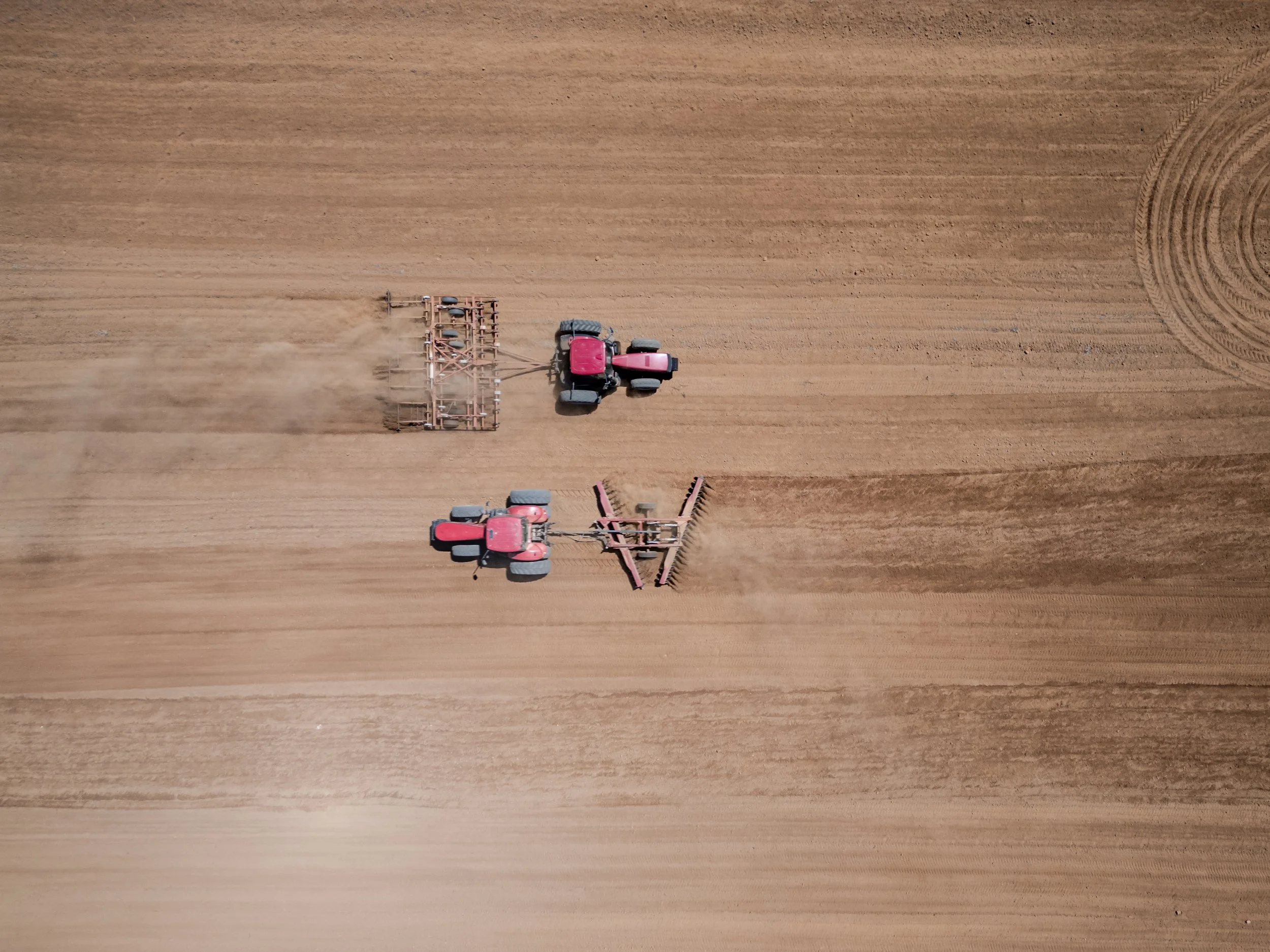 An aerial view of a red tractor pulling a cultivator across a large, flat, brown field with visible tire tracks on the right side.