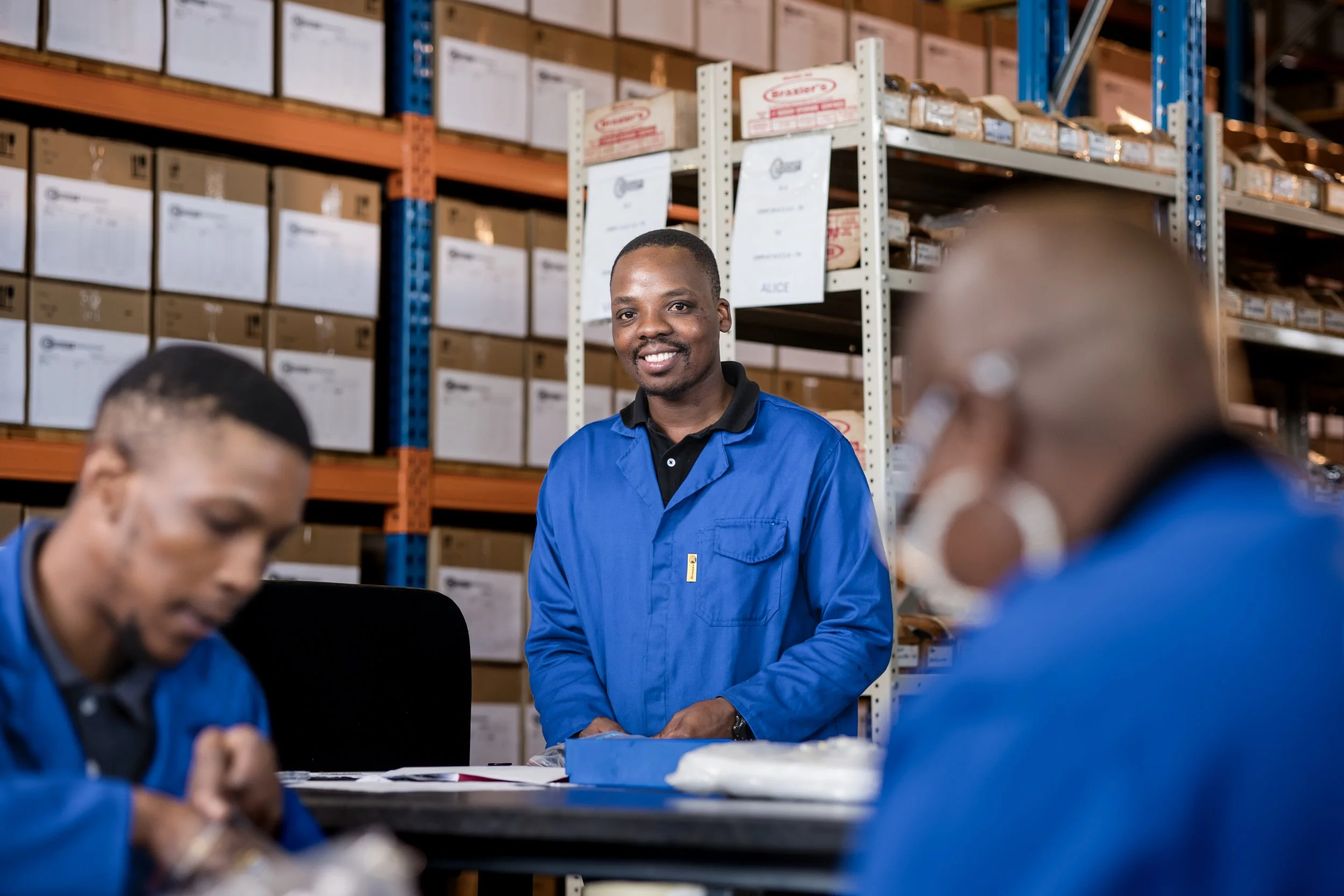 A man in a blue uniform smiling in a warehouse with shelves filled with boxes. Other workers are in the foreground.