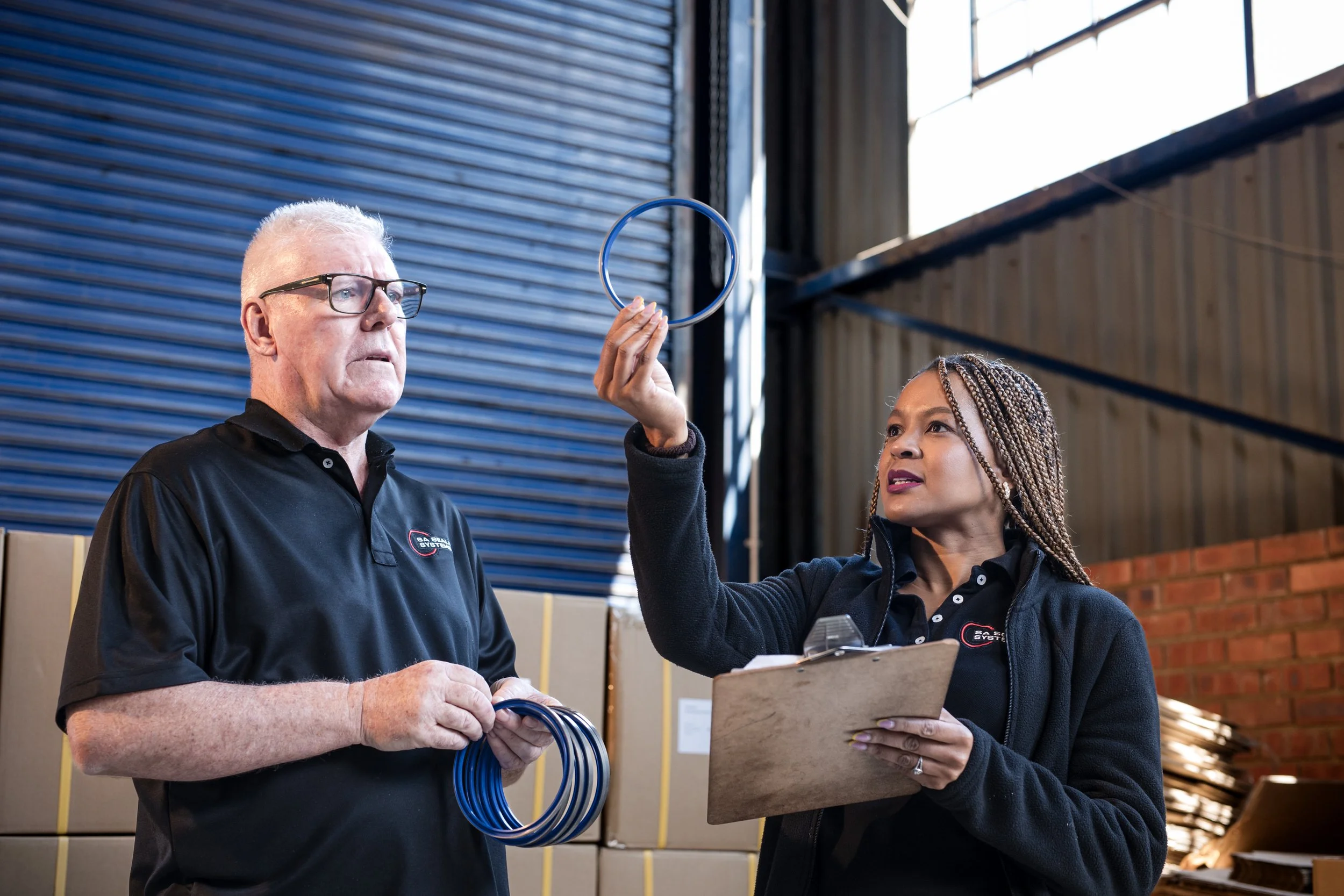 Two workers in a warehouse, one man and one woman, inspecting seals, with the woman holding a clipboard and information, and the man holding a bundle of seals, against a background of metal doors and a brick wall.