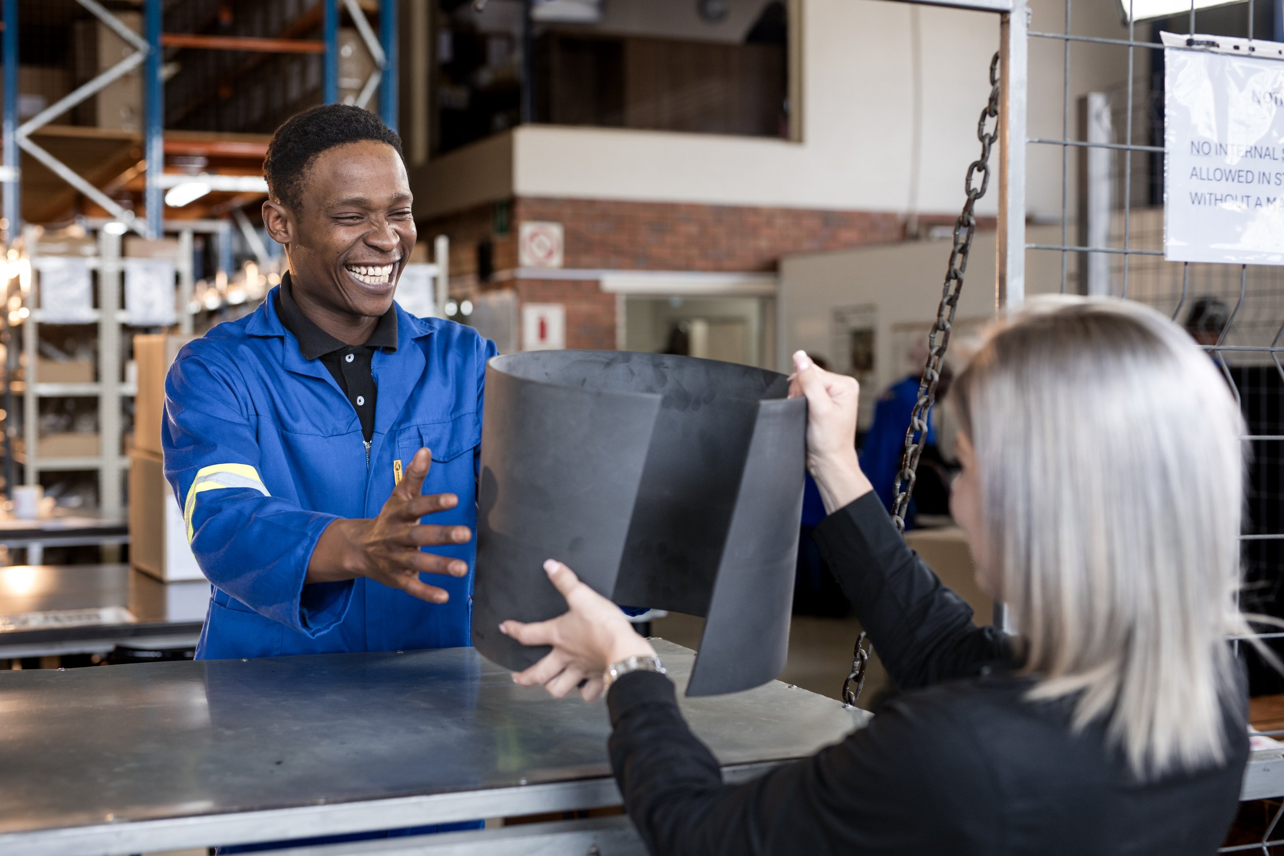 A smiling workplace employee in a blue uniform handing a large black planter to a customer at a store counter.