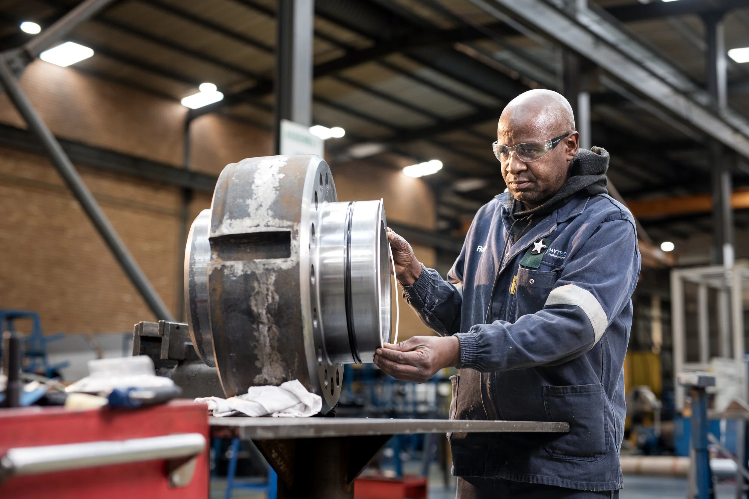 A robotic or machine part is being examined by a man in a factory or workshop. The man, wearing safety glasses and work clothing, is closely inspecting the metal component.