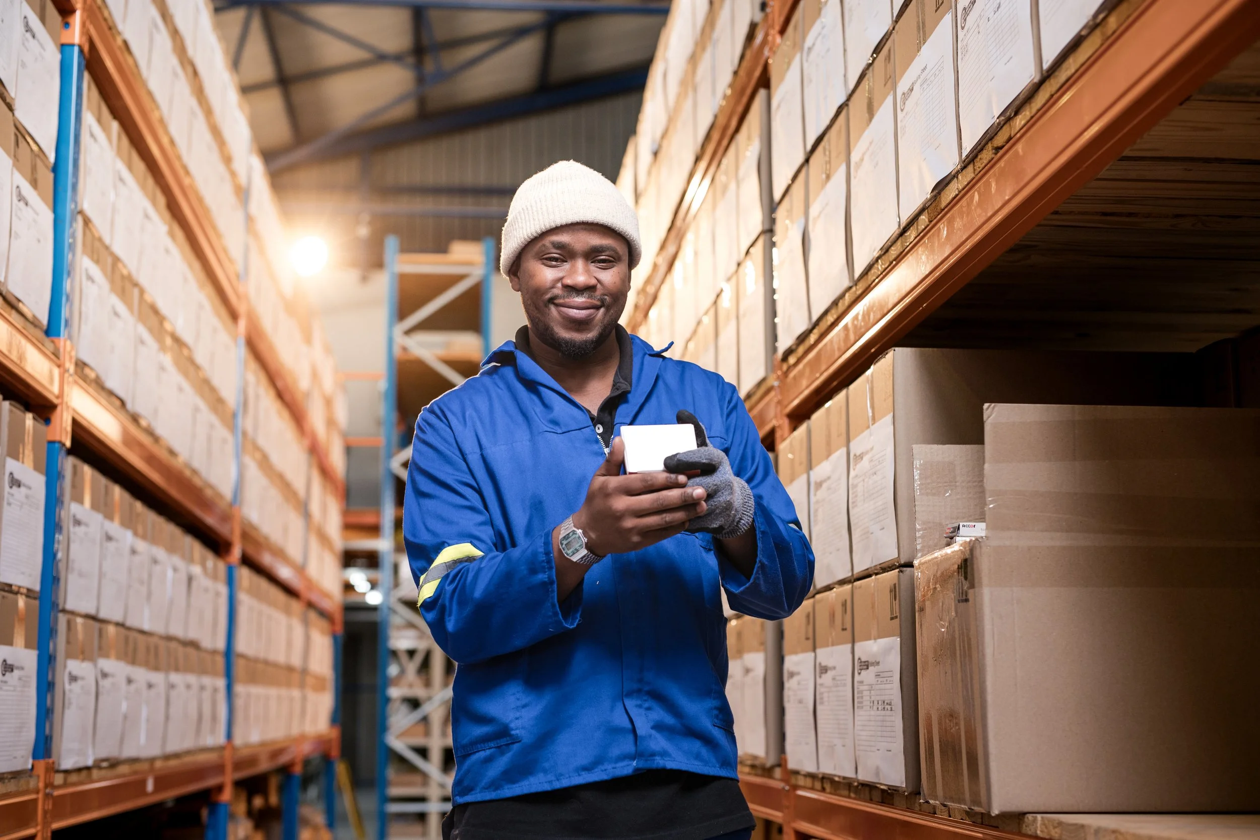 A warehouse worker in a blue uniform and white beanie, smiling, holding a device, standing among stacked cardboard boxes on orange shelving.