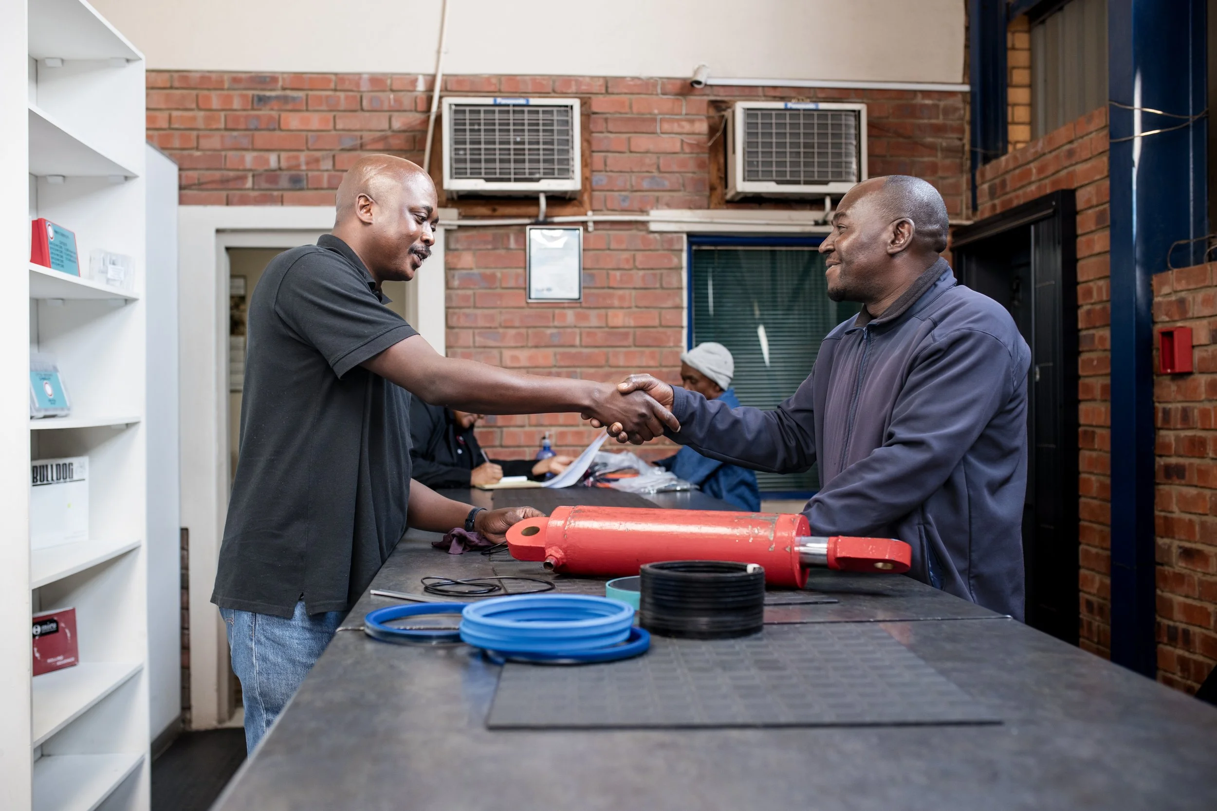 Two men shaking hands inside a workshop with brick walls and air conditioning units on the wall.