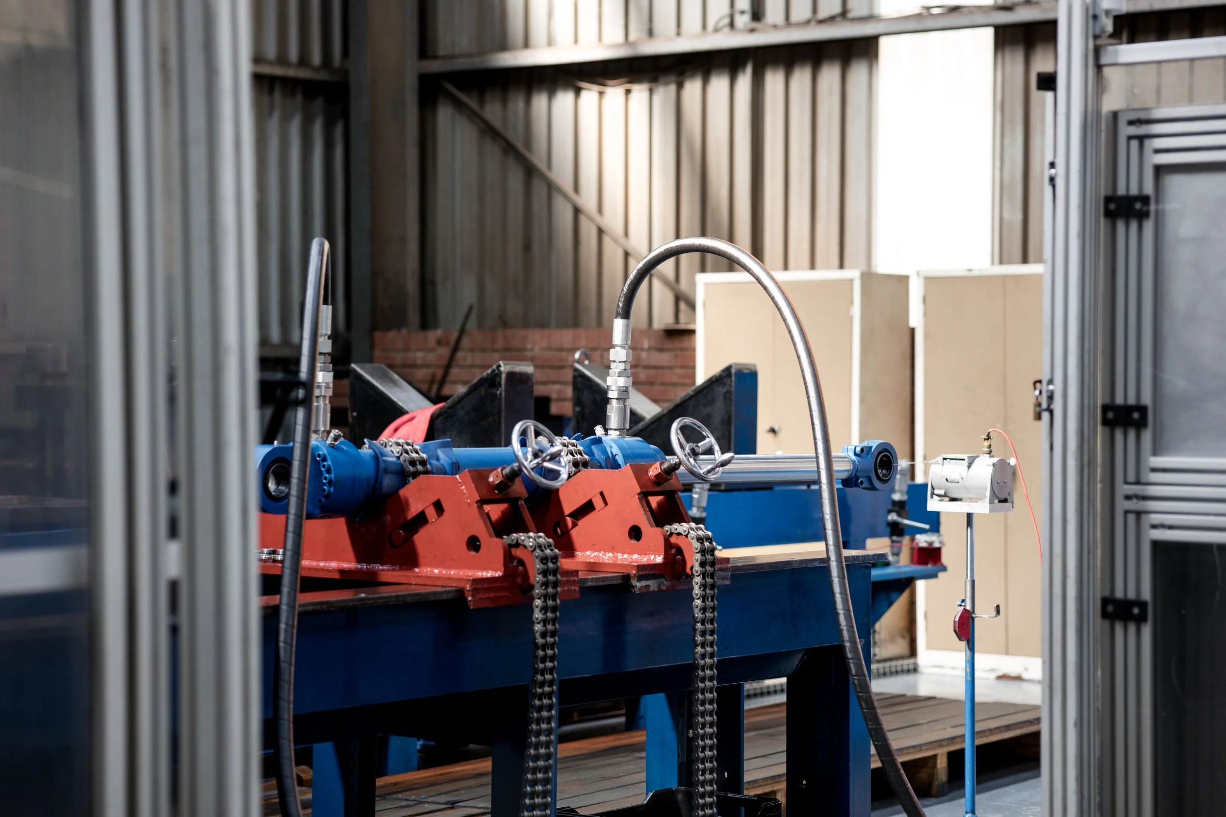 Industrial machinery with blue and red components, chains, and pipes, inside a warehouse or factory with metal siding and wooden elements in the background.