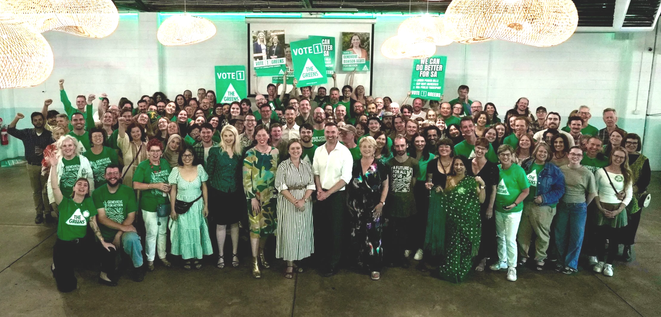 A large group of people gathered indoors at a political event supporting The Greens. Many are smiling, some raising clenched fists, and some holding signs with campaign slogans. The crowd is diverse, with several people wearing green shirts or clothing indicating support for The Greens.