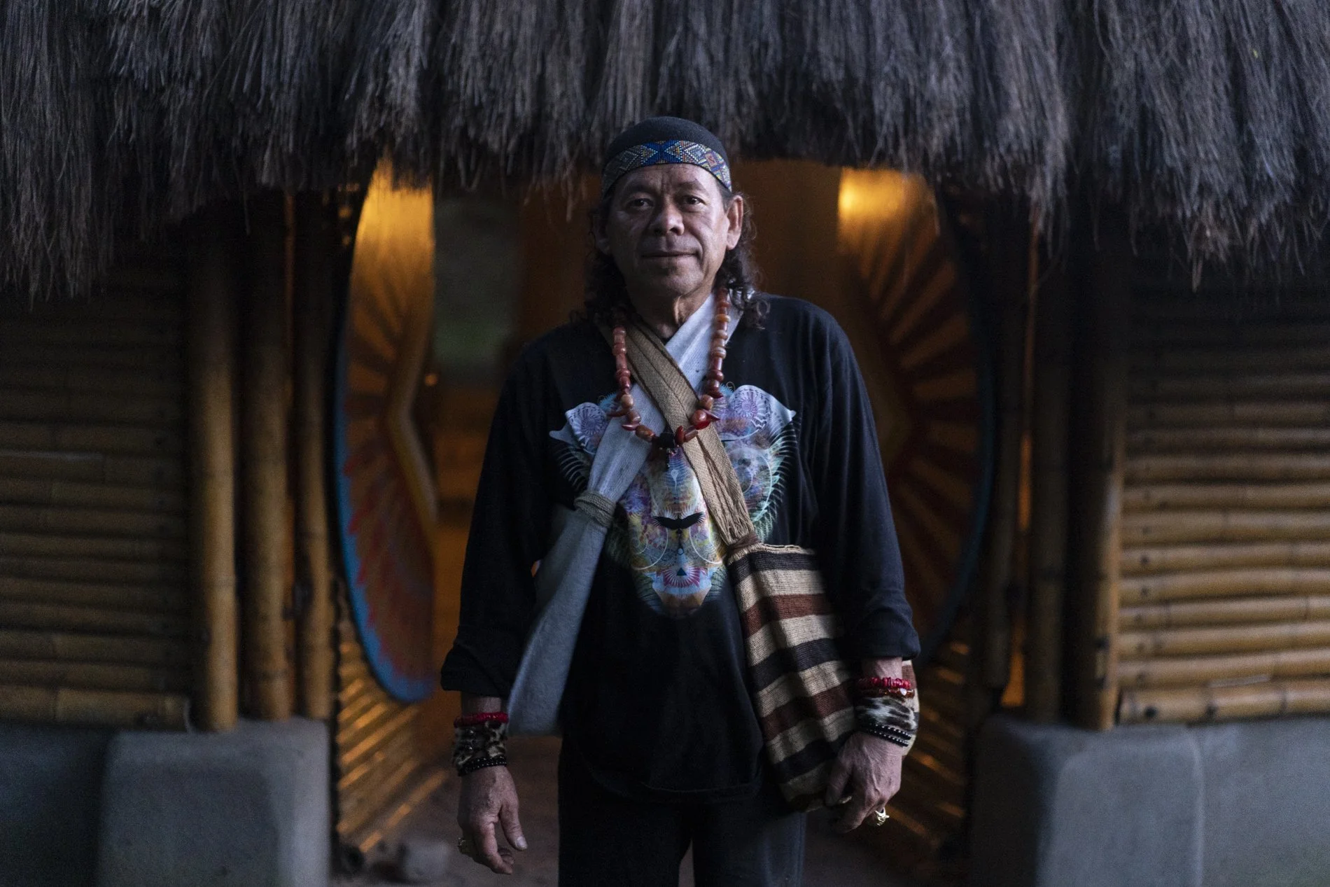 A man in traditional indigenous clothing stands in front of a bamboo and straw hut with an open doorway, during daytime or dusk.