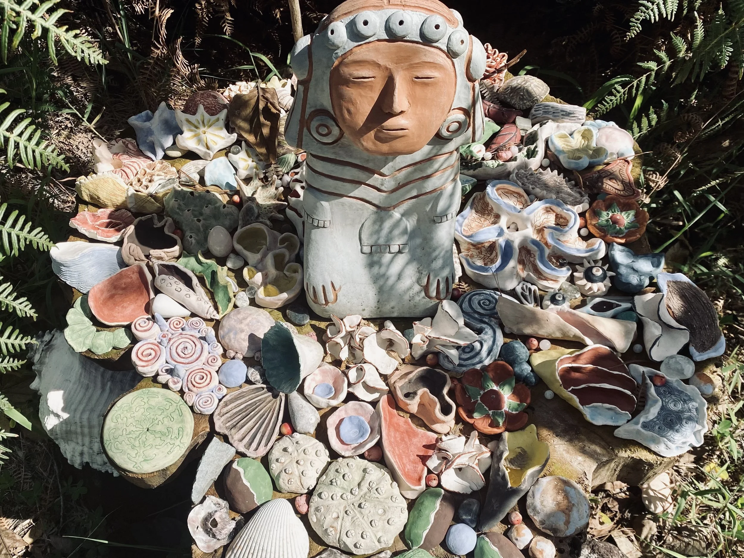 A sculpture of a human face with closed eyes, surrounded by various colorful seashells and marine-themed decorative items, on a natural outdoor setting with ferns and foliage in the background.