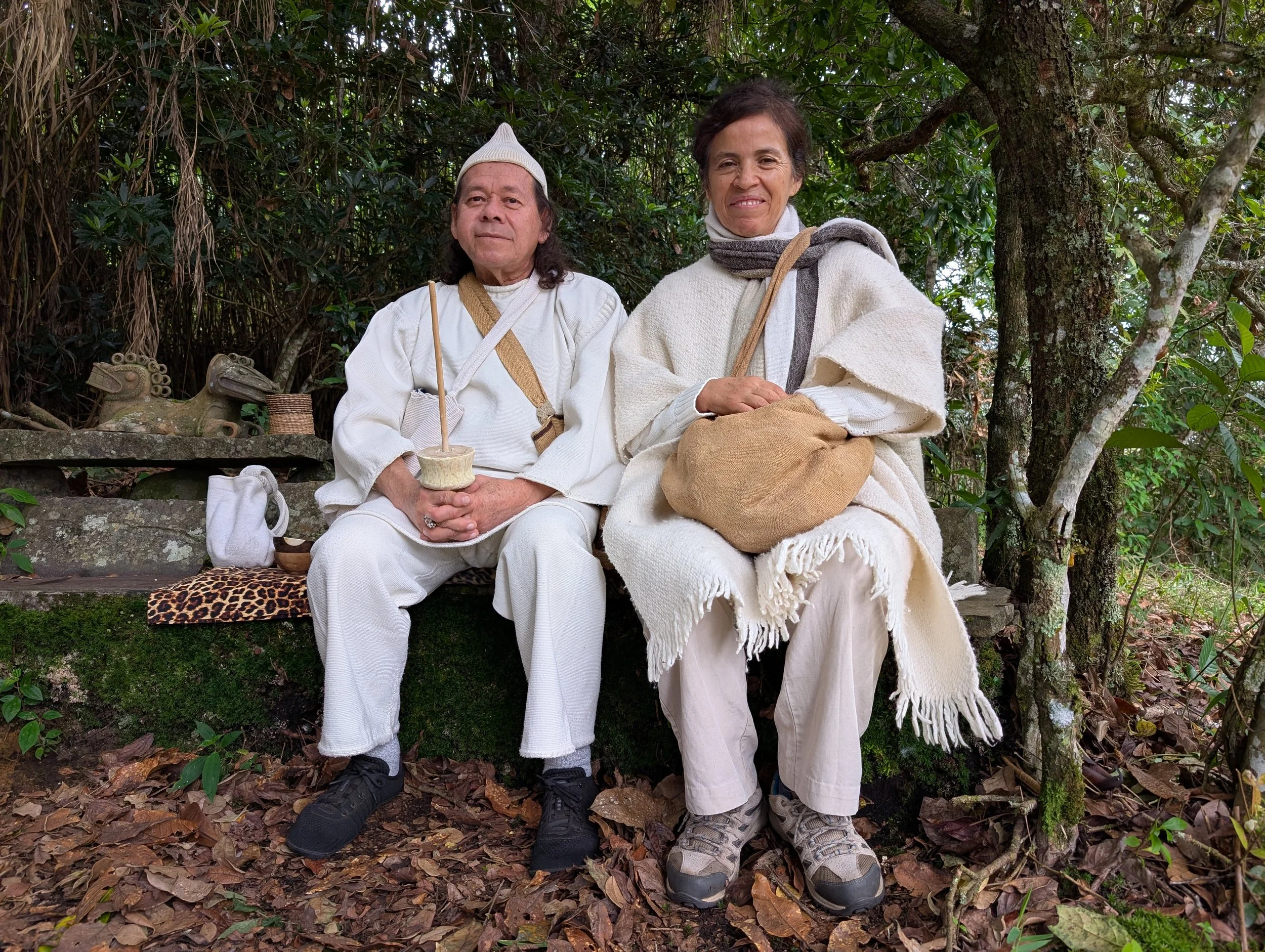 Two people in white clothing sitting outdoors in a forested area. The man on the left has long hair, a white hat, and is holding a small ceramic bowl. The woman on the right has short hair, is smiling, and is wearing a white shawl and scarf, with a burlap sack on her lap. They are surrounded by trees and leaves, with traditional artifacts, including small animal sculptures, nearby.
