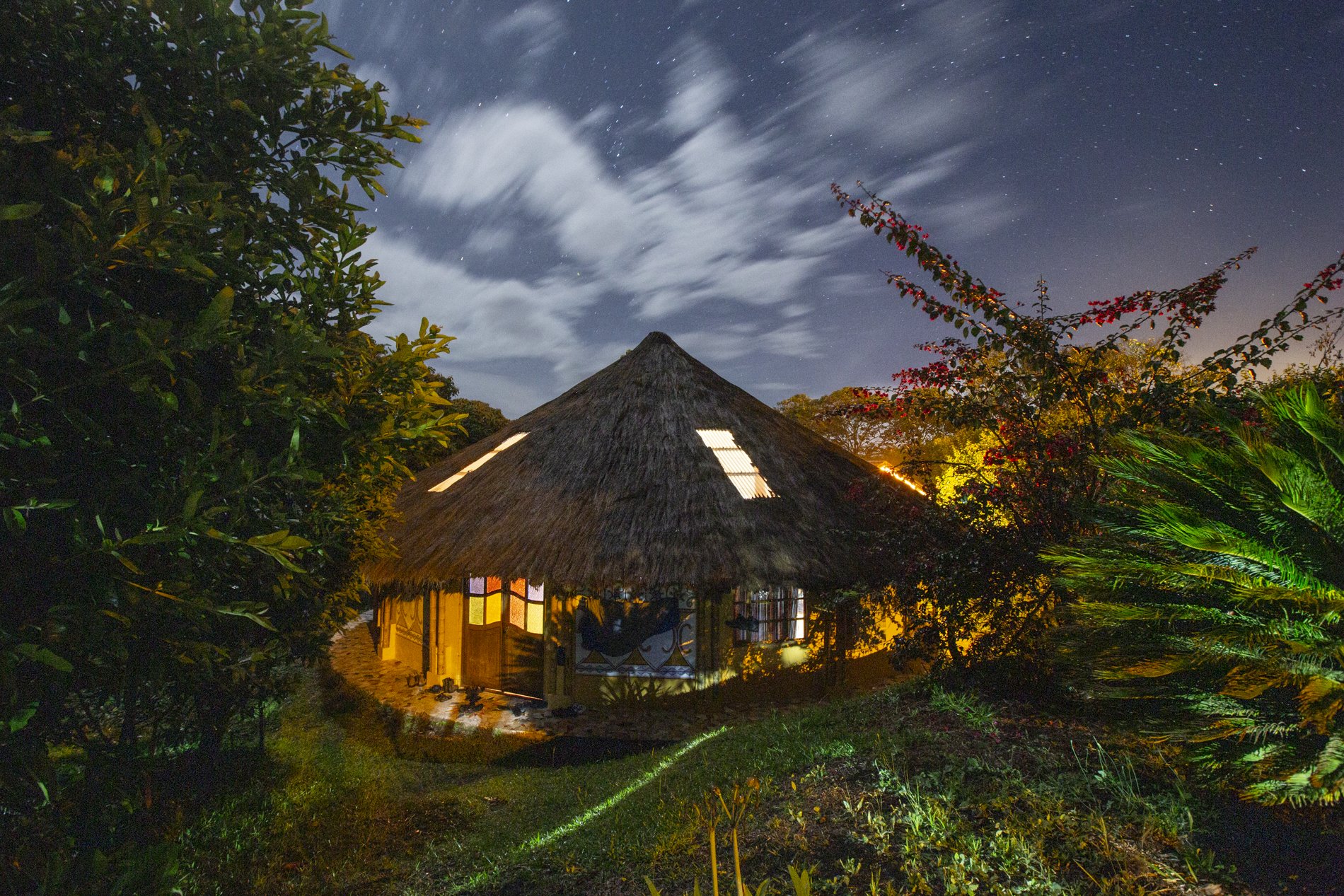 A cozy thatched-roof hut illuminated from within, surrounded by lush greenery and colorful plants, under a starry night sky with moving clouds.