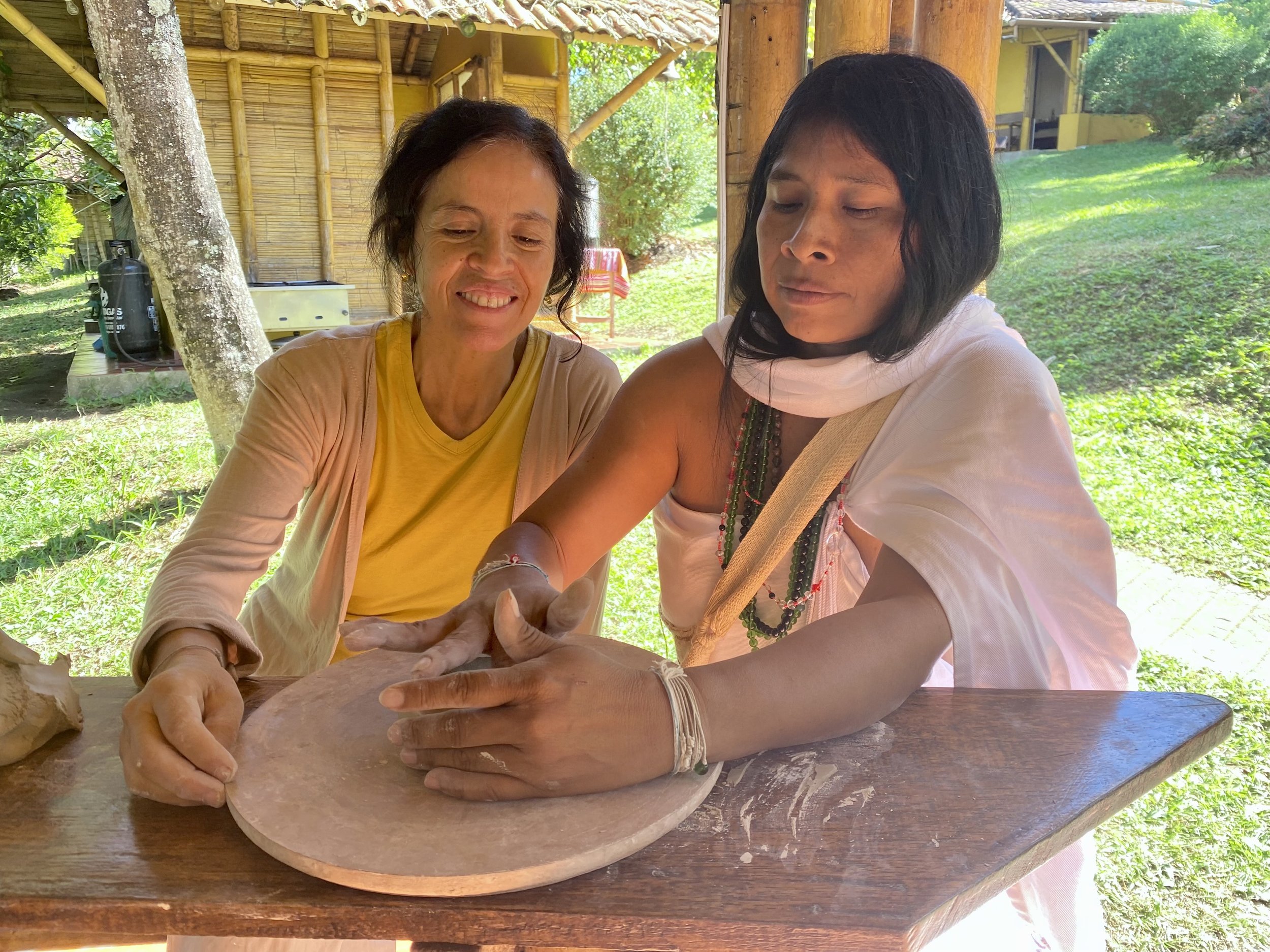Two women, one older and one younger, are sitting at a wooden table outdoors, rolling out some dough together. The background shows greenery, trees, and a rustic bamboo and wooden structure.