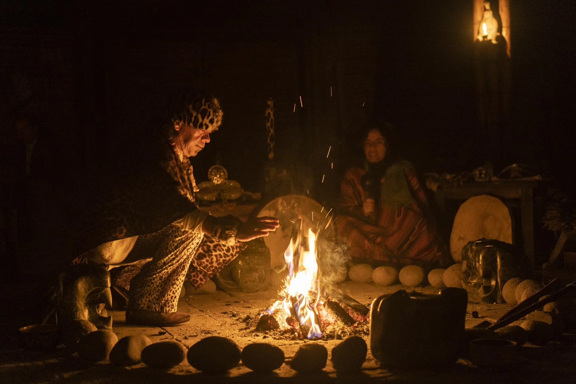 Two people sitting by a campfire at night, one wearing leopard print clothing and the other wearing a colorful striped poncho, with a lit candle inside a framed opening in the background.