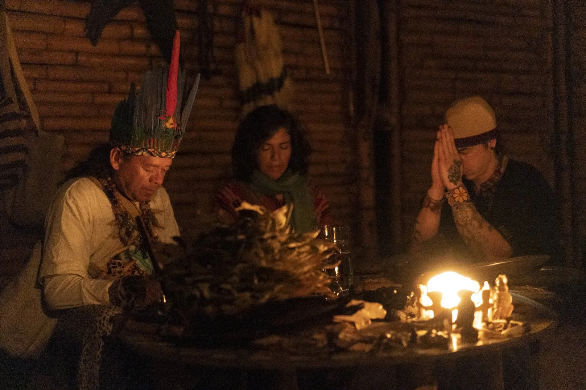 Three people sitting around a table illuminated by candlelight, with one person wearing an elaborate feathered headdress and traditional attire, another person with curly dark hair and a scarf, and the third person with tattoos on their arms and a beige cap with glasses, in a dimly lit wooden interior.