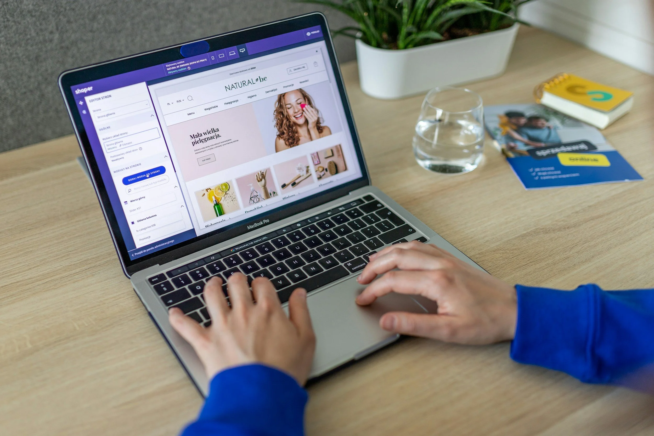 Person using a MacBook Pro laptop on a wooden desk, viewing an online store webpage, with a glass of water, magazines, and a potted plant nearby.