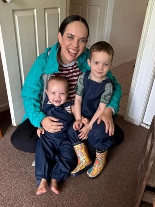 A woman with dark hair smiling, sitting on the floor with two young children, all dressed in rain gear. The children are wearing navy rain suits, and the woman is wearing a bright blue jacket. They are indoors near a door, with a cardboard box in front of them.
