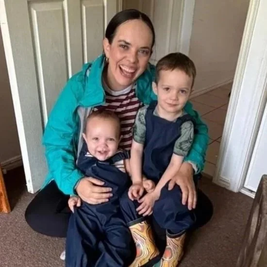 A woman sitting on the floor with two young boys. She is smiling, wearing a bright blue jacket over a striped shirt. The younger boy has Down syndrome and is sitting on her lap, smiling widely. The older boy is sitting next to her with a neutral expression, wearing a dark vest and patterned rain boots. They are indoors, next to a door.