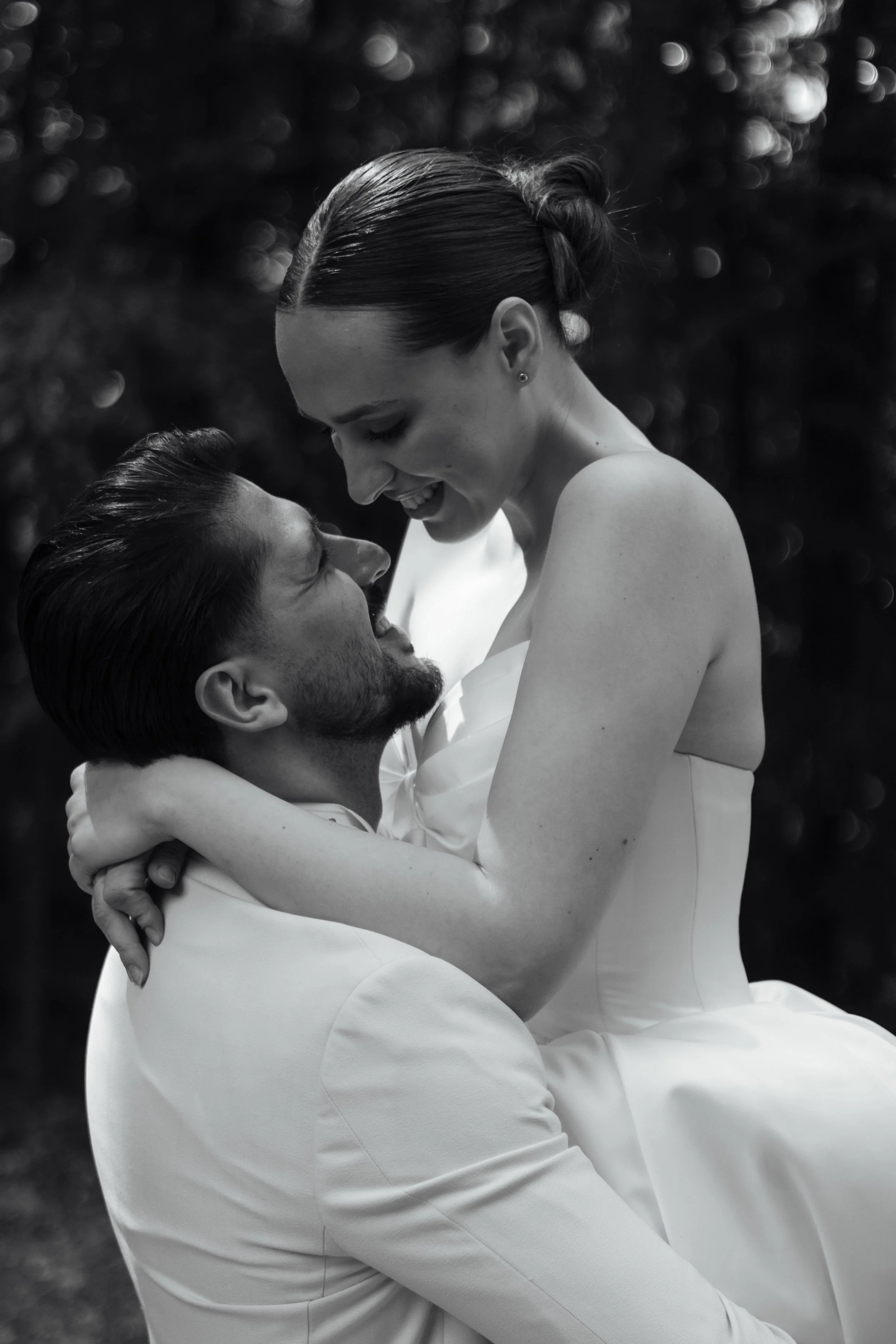 Ein schwarz-weiß Foto eines Paares, das sich liebt. Die Frau sitzt auf den Beinen des Mannes, beide schauen sich an und lächeln. Der Hintergrund ist unscharf mit Bokeh-Effekt.