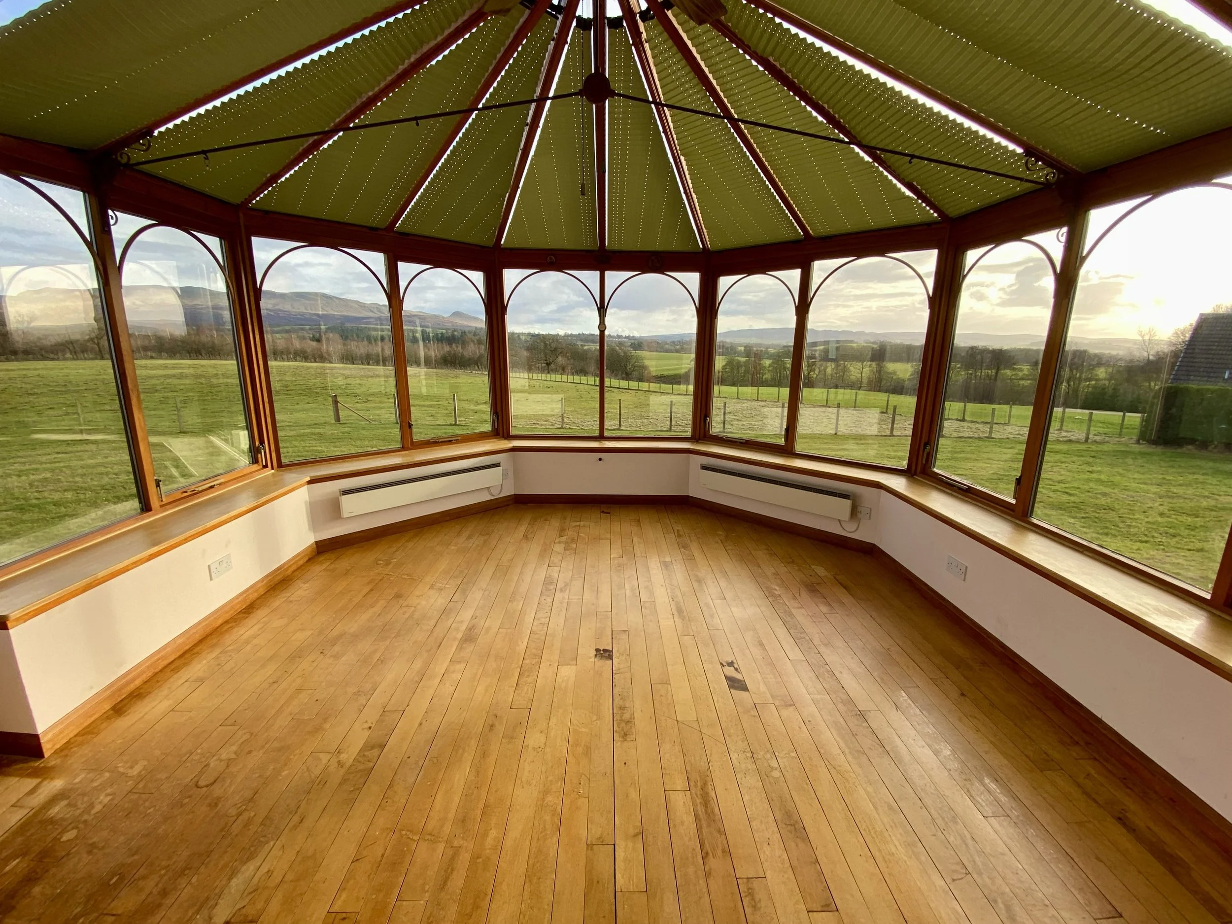 Interior of a sunroom with large glass windows overlooking a green landscape and mountains in the background, featuring a circular design and wooden flooring.