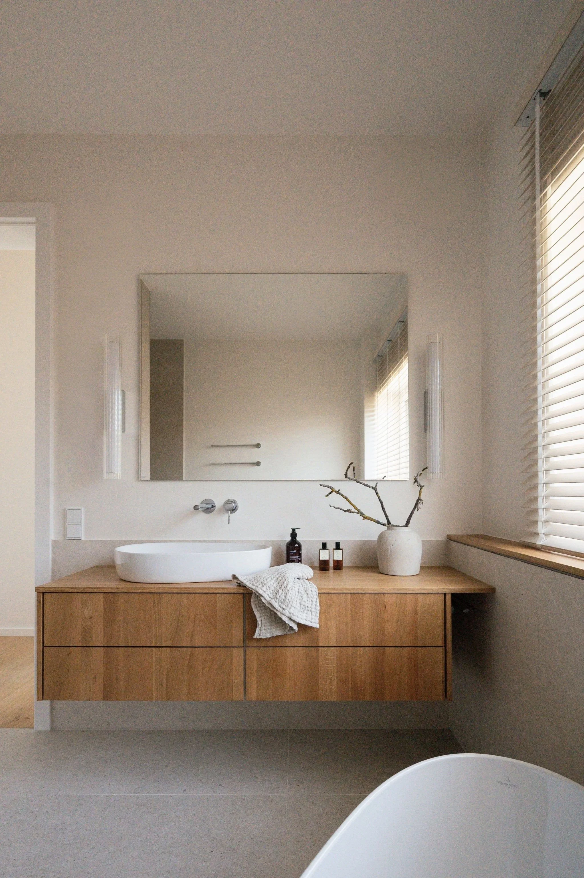 Ein modernes Badezimmer mit einem Waschtisch aus Holz, einem ovalen weißen Waschbecken, einem großen Spiegel, Vasen mit Zweigen, Handtüchern, Lotionflaschen und einem Fenster mit Jalousien.