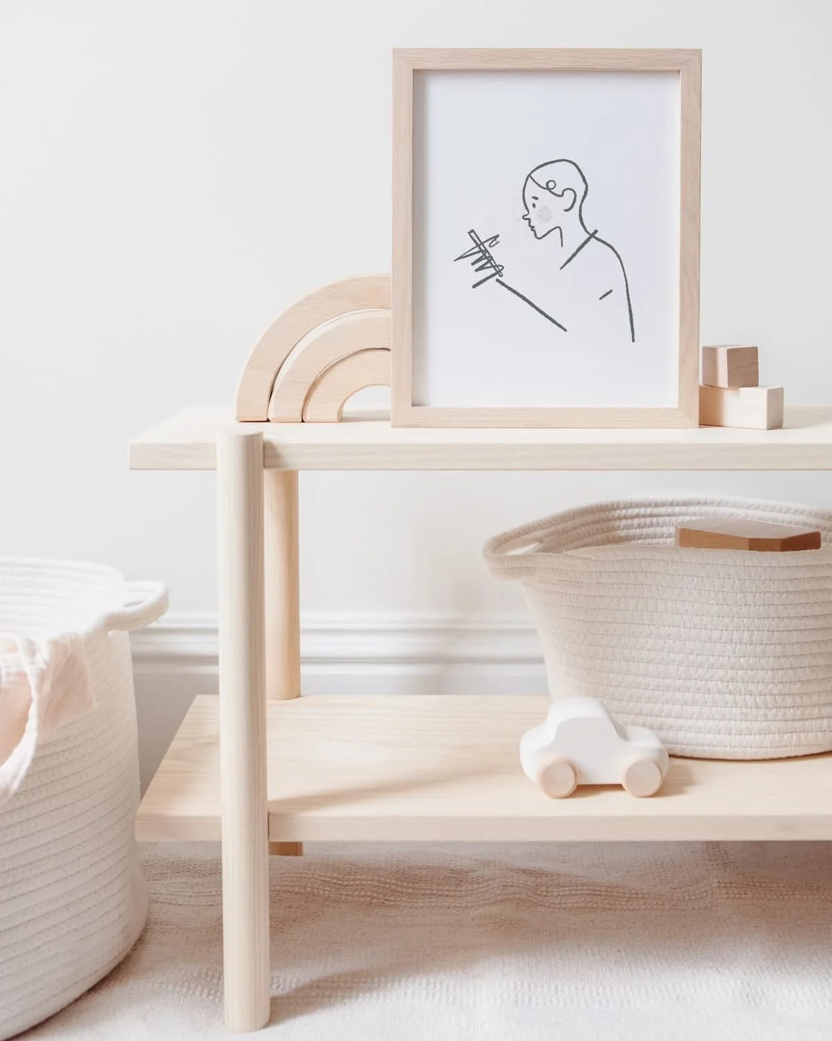 Minimalistisches Kinderspielzeug und Dekoration in hellen, natürlichen Tönen, einschließlich Korb, Holzauto, Bogen und Bilderrahmen mit Strichzeichnung eines Kindes mit Spritze.