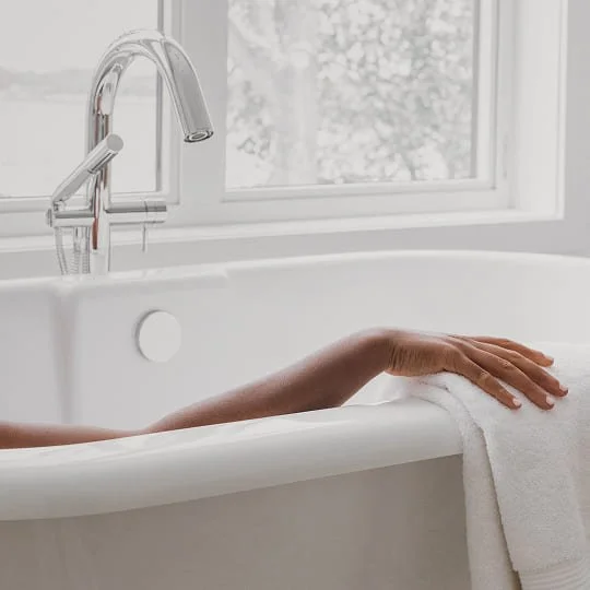 Eine Frauenhand liegt entspannt auf dem Rand einer Badewanne vor einem Fenster