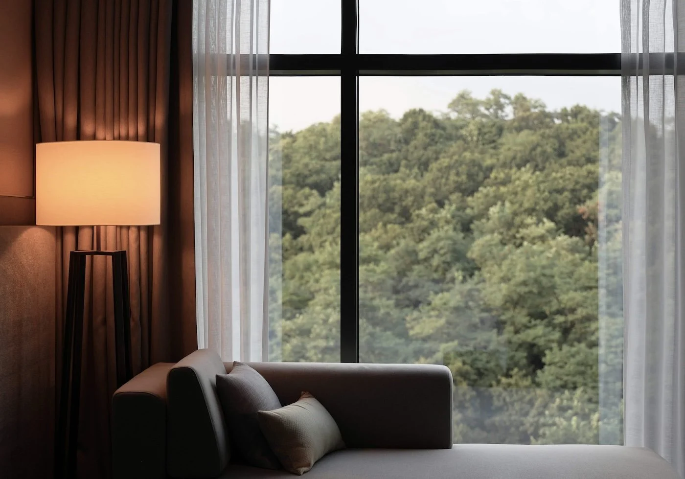 Das Foto zeigt einen hotelähnlichen Raum mit Blick ins Grüne, einer bequemen Sitzgelegenheit und einer leuchte mit warmen Licht