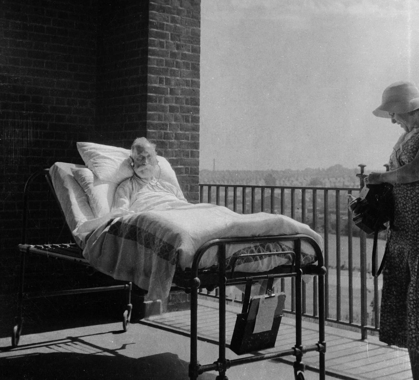 Das Foto zeigt einen alten kranken Mann im Krankenhaus. Man hat ihn mit dem Krankenbett auf einen Balkon geschoben, damit er Sonne und frische Luft bekommt