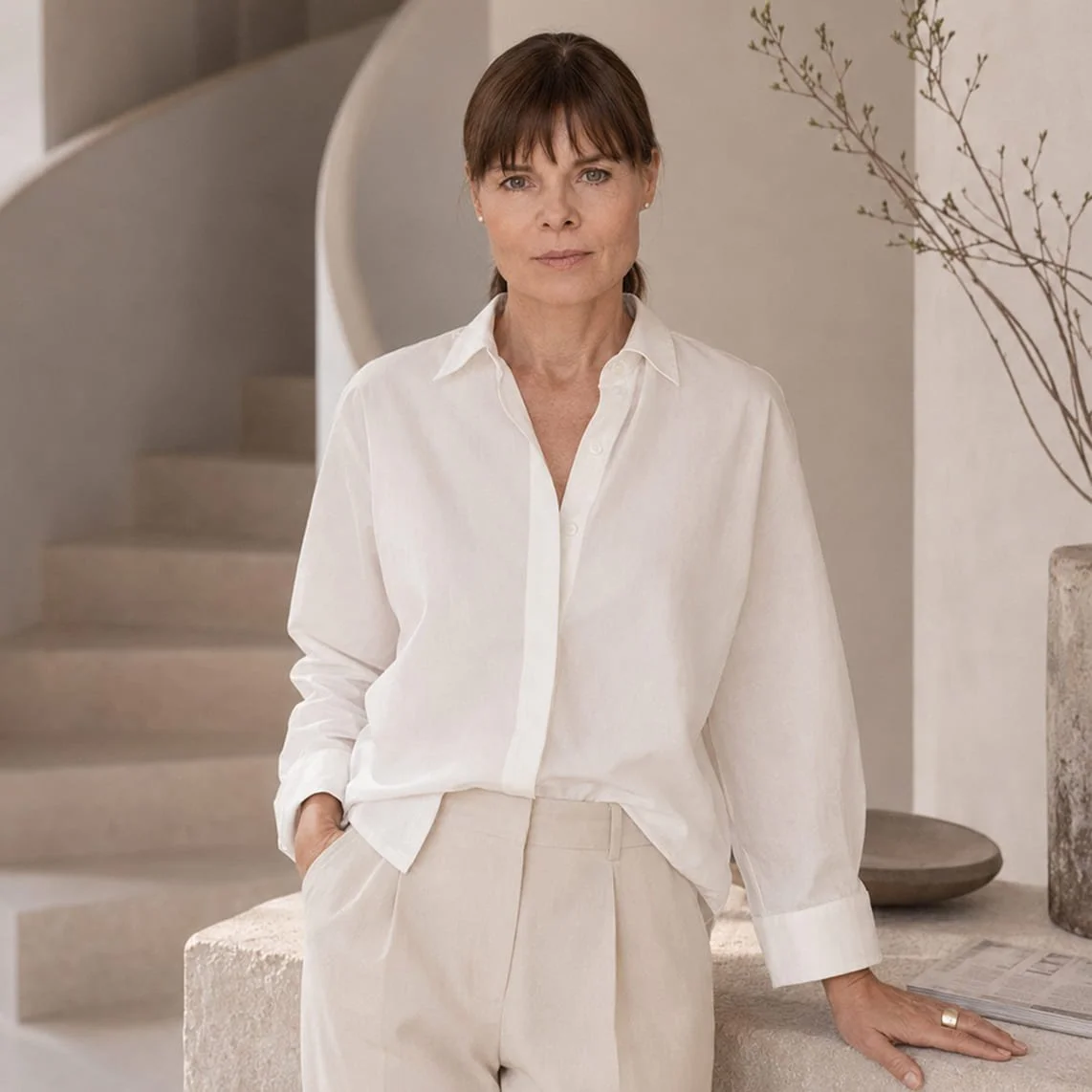 Martina Velmeden in weißem Hemd und beigen Hosen lehnt an einer Stelle in einem minimalistischen Raum mit Treppe und Dekorationen aus Naturmaterialien.
