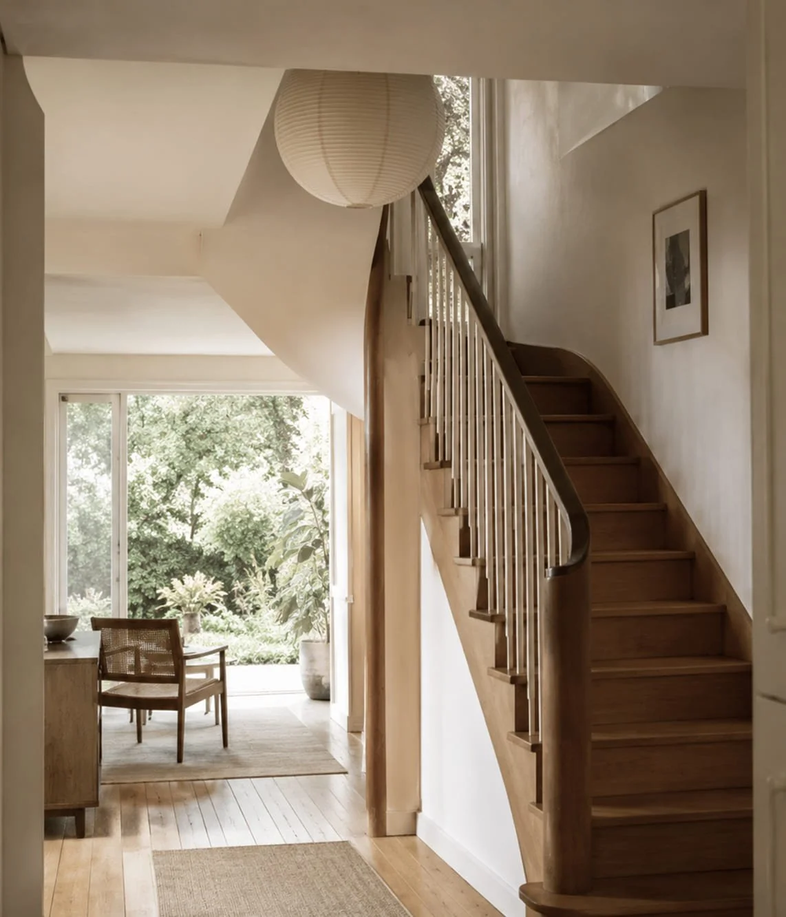 Innenansicht eines Hauses mit einer Holztreppe, großen Fenster, einem Stuhl und Pflanzen im Hintergrund.
