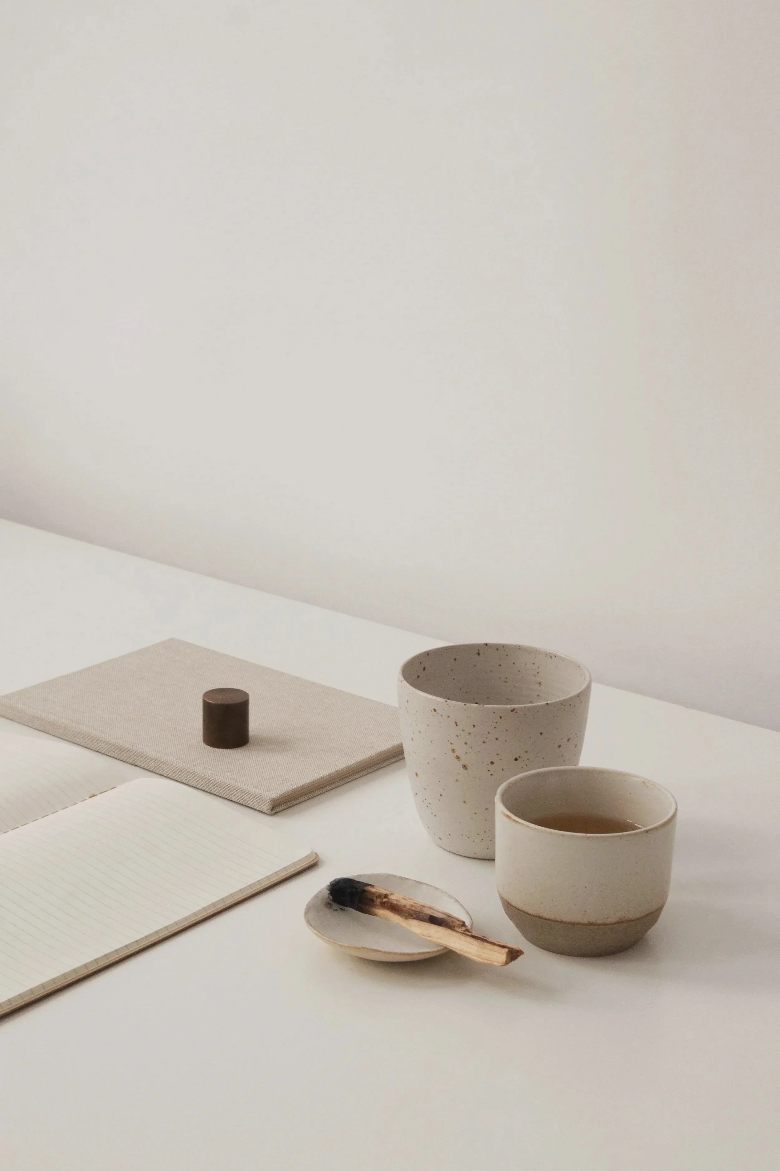 Weißer Tisch mit zwei gepflasterten Tassen, einem kleinen Teller mit Holzlöffel, Notizbüchern, einem Stift, Kerzen und einer beige Tischmatte. Hintergrund: weiße Wand, gedeckter Raumstil.