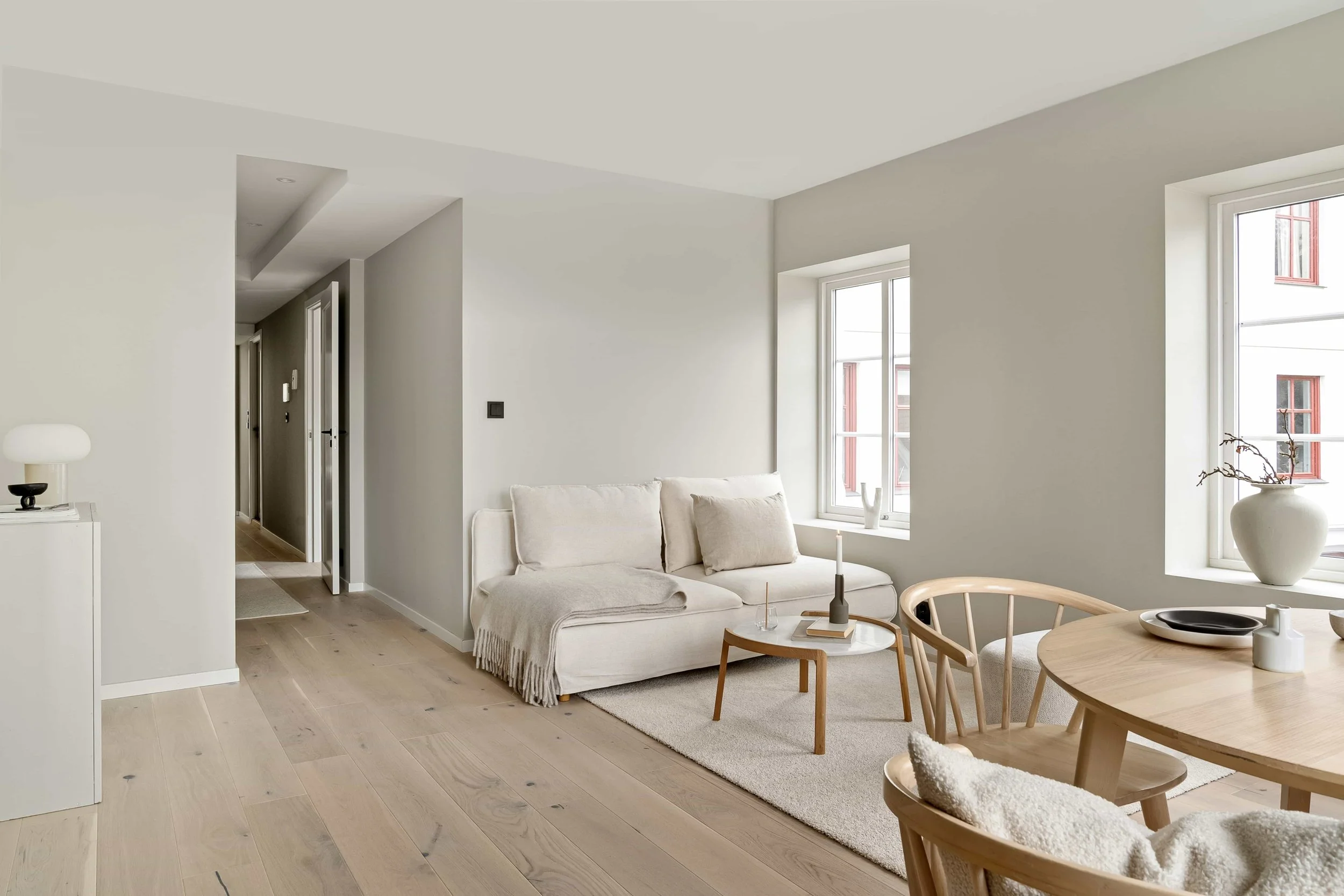 Helles Wohnzimmer mit großen Fenstern, weißen Wänden, beige Couch, Teetisch, Holzstuhl und Dekorationsgegenständen