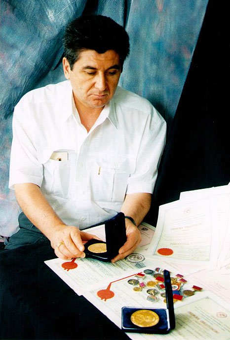 Man in a white shirt looking at a medal in a display case, with documents and scattered coins and medals on a table.