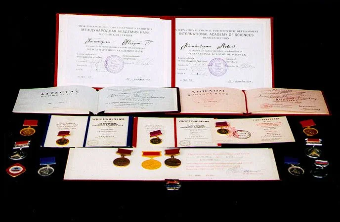 Display of international awards, medals, and certificates on a black background, including diplomas from the International Academy of Sciences with signatures and stamps.