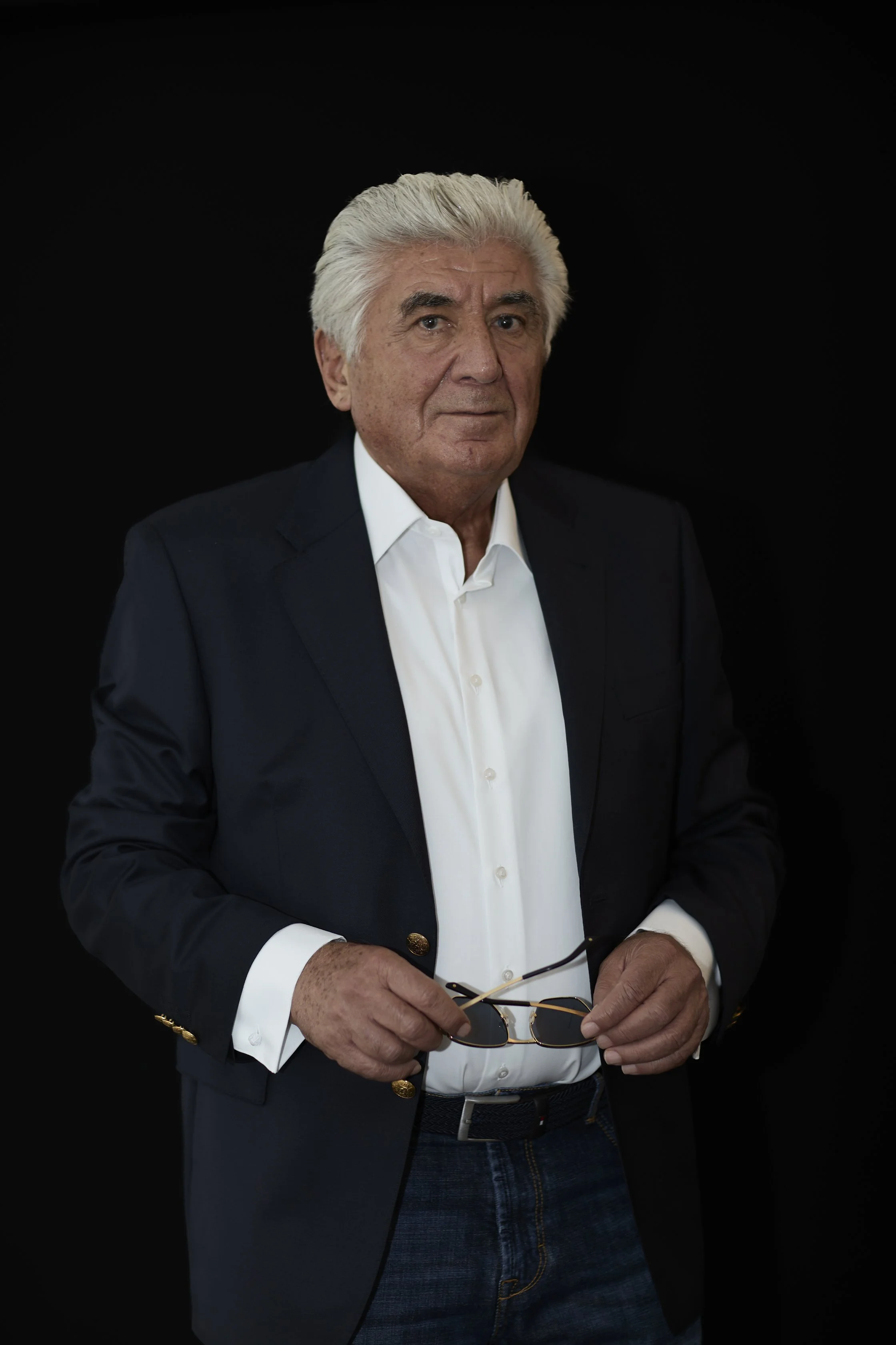 A man with gray hair wearing a dark suit and white shirt, holding sunglasses in front of a black background.