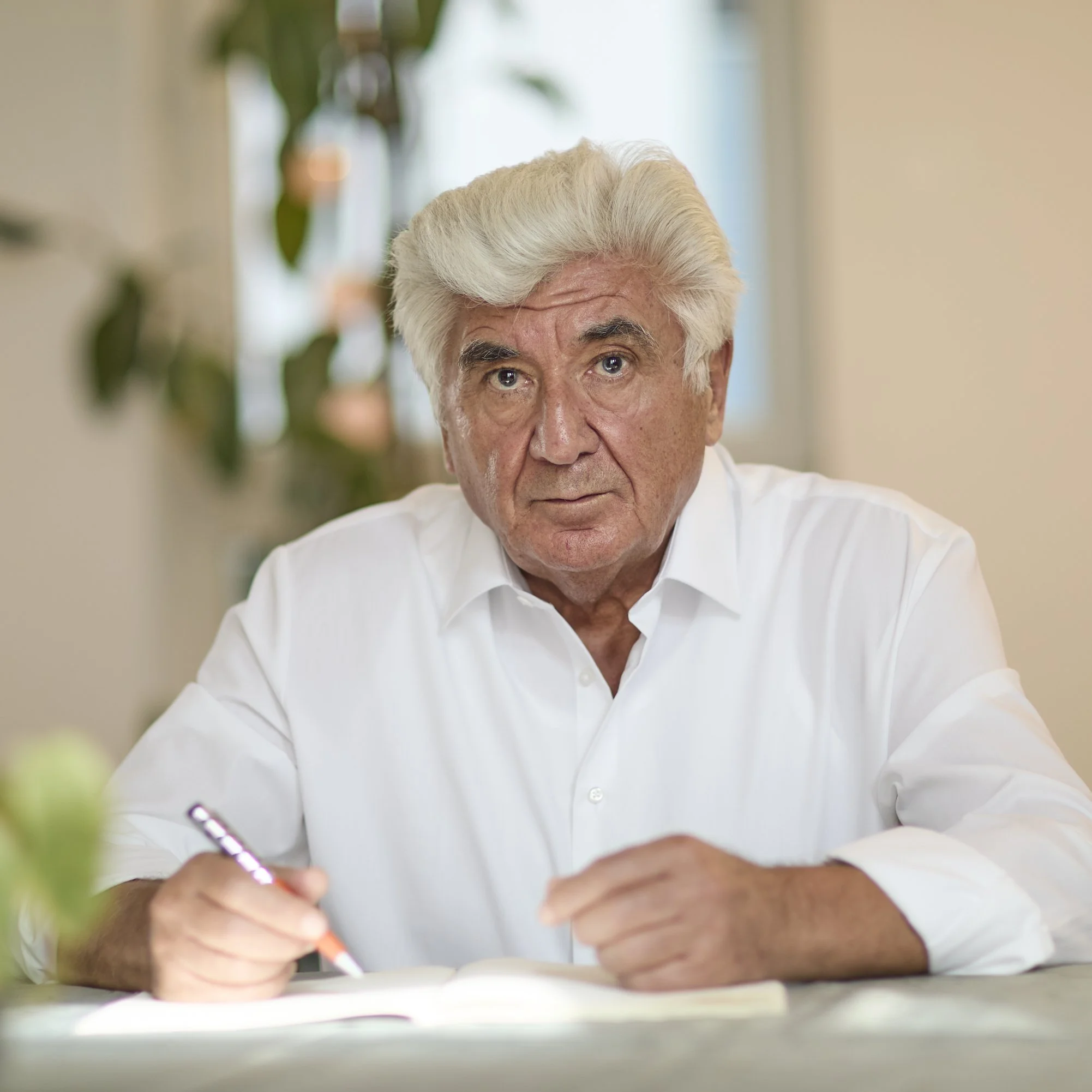 The photo is taken from a low angle, with the man wearing a white shirt. The background is blurred, showing some green leaves and beige walls.