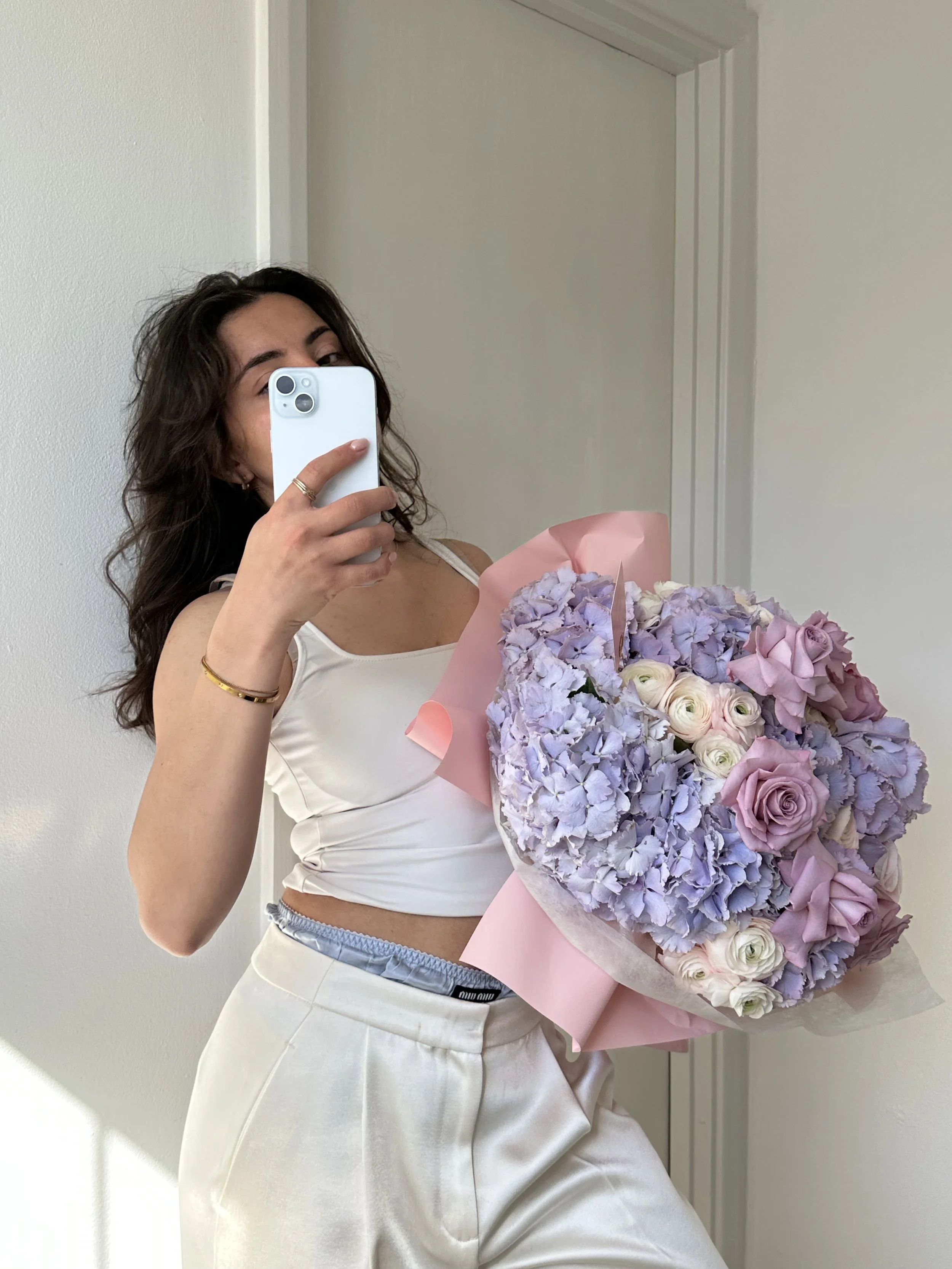 A woman with dark, wavy hair taking a mirror selfie with a smartphone. She is holding a large bouquet of pastel-colored flowers, including hydrangeas and roses wrapped in pink paper. She is wearing a white, sleeveless top and white pants with a silver belt.
