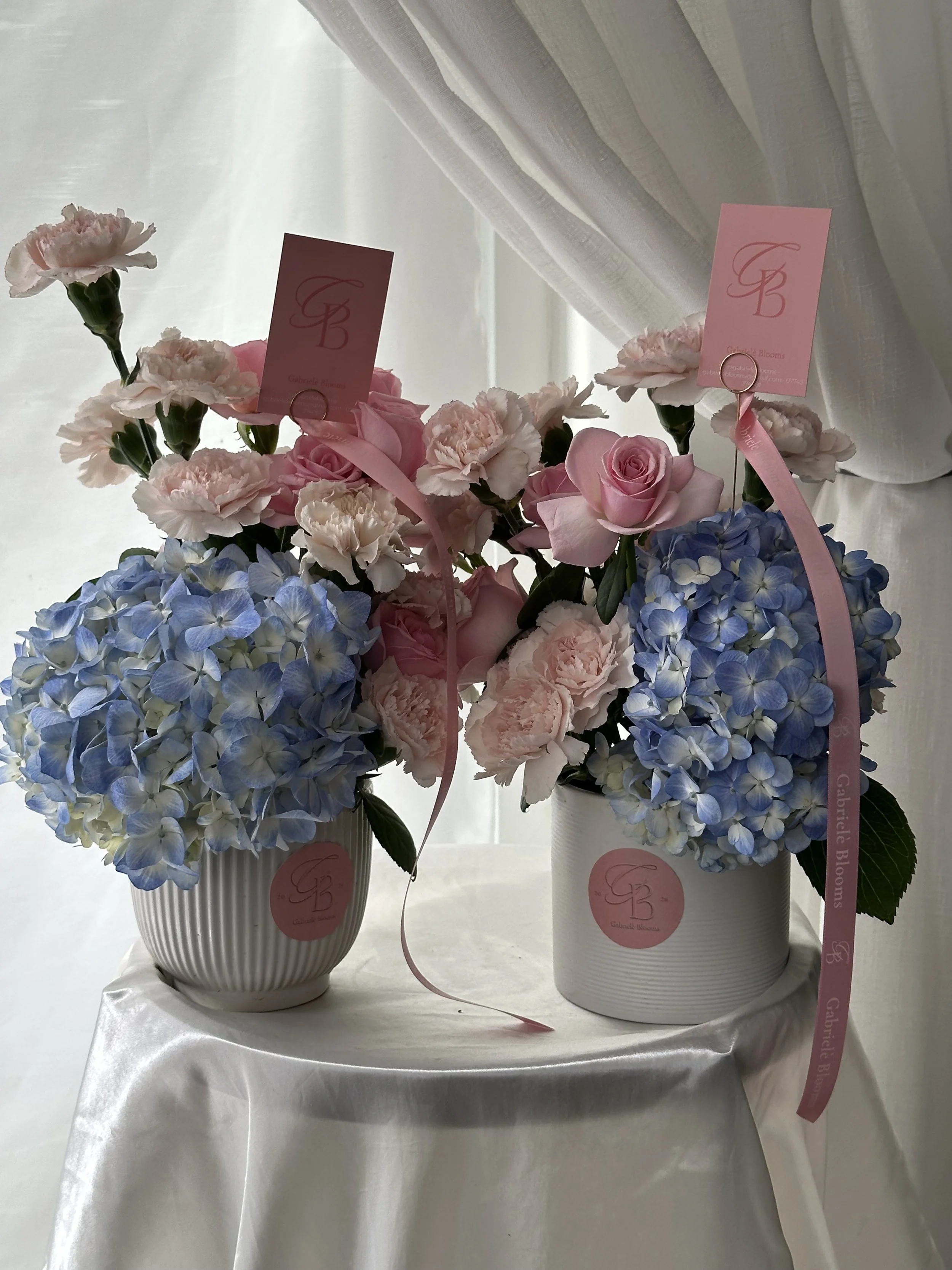 Two white vases with pink and blue flowers, labeled with pink tags featuring a logo and the text 'Gabrielle Blooms' on pink ribbons, set on a white tablecloth with a sheer white curtain in the background.