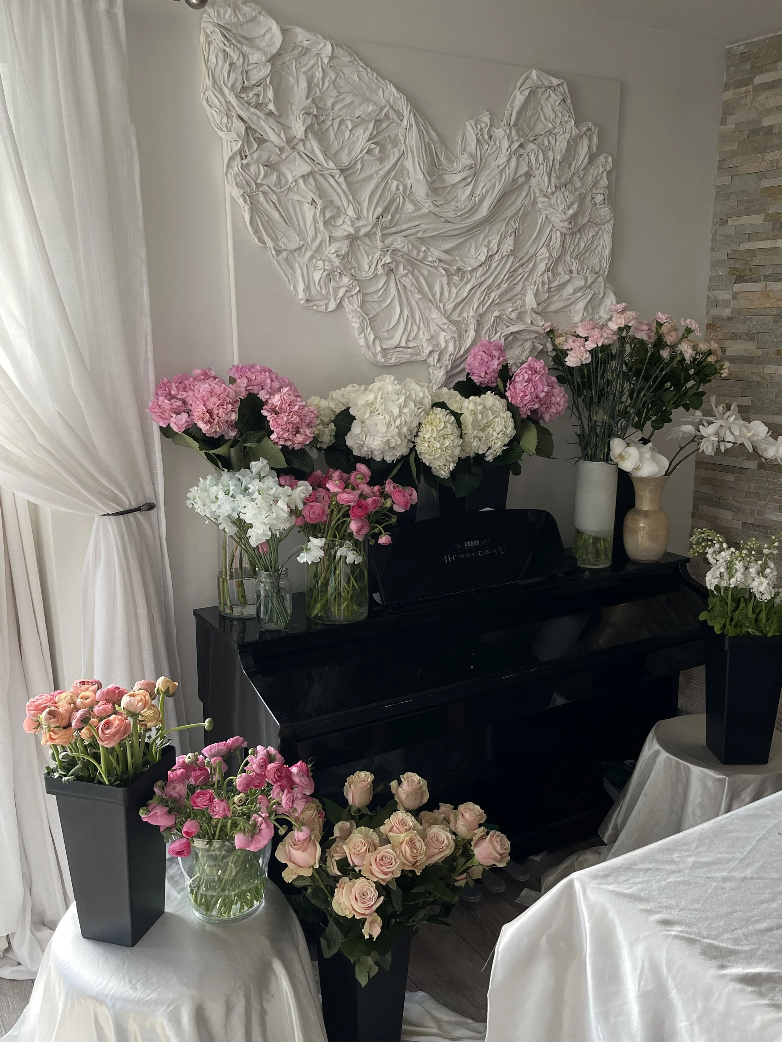 A black piano with prepped flowers ready to arrange of pink, white, and cream roses and hydrangeas in vases on top and around it, set in a room with cream-colored drapes, a textured wall, and a draped white cloth.