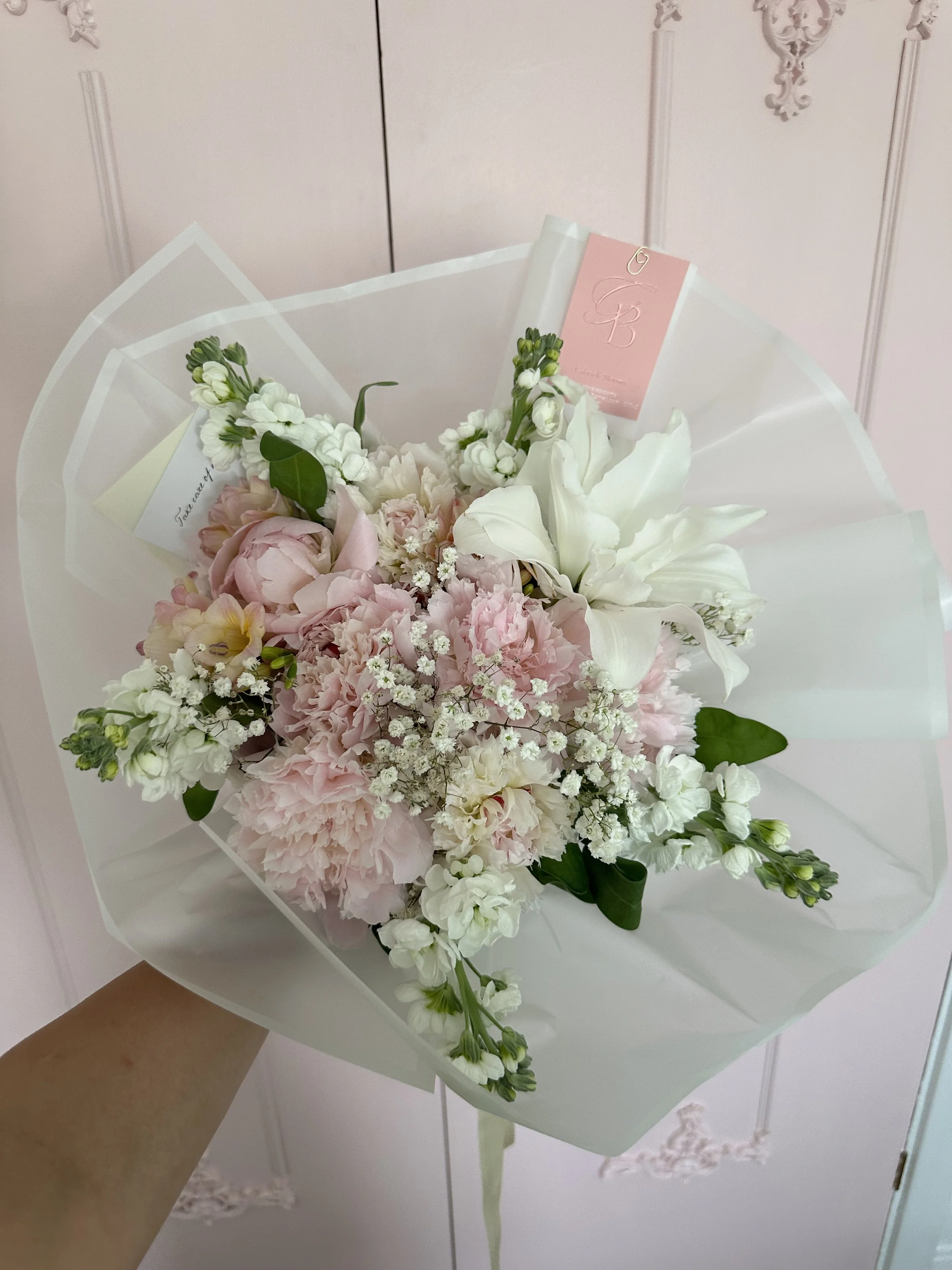 A bridal bouquet featuring pink peonies, white lilies, white roses, and small white filler flowers, wrapped in white paper with a pink and white tag, held by a person's hand against a pink wall.