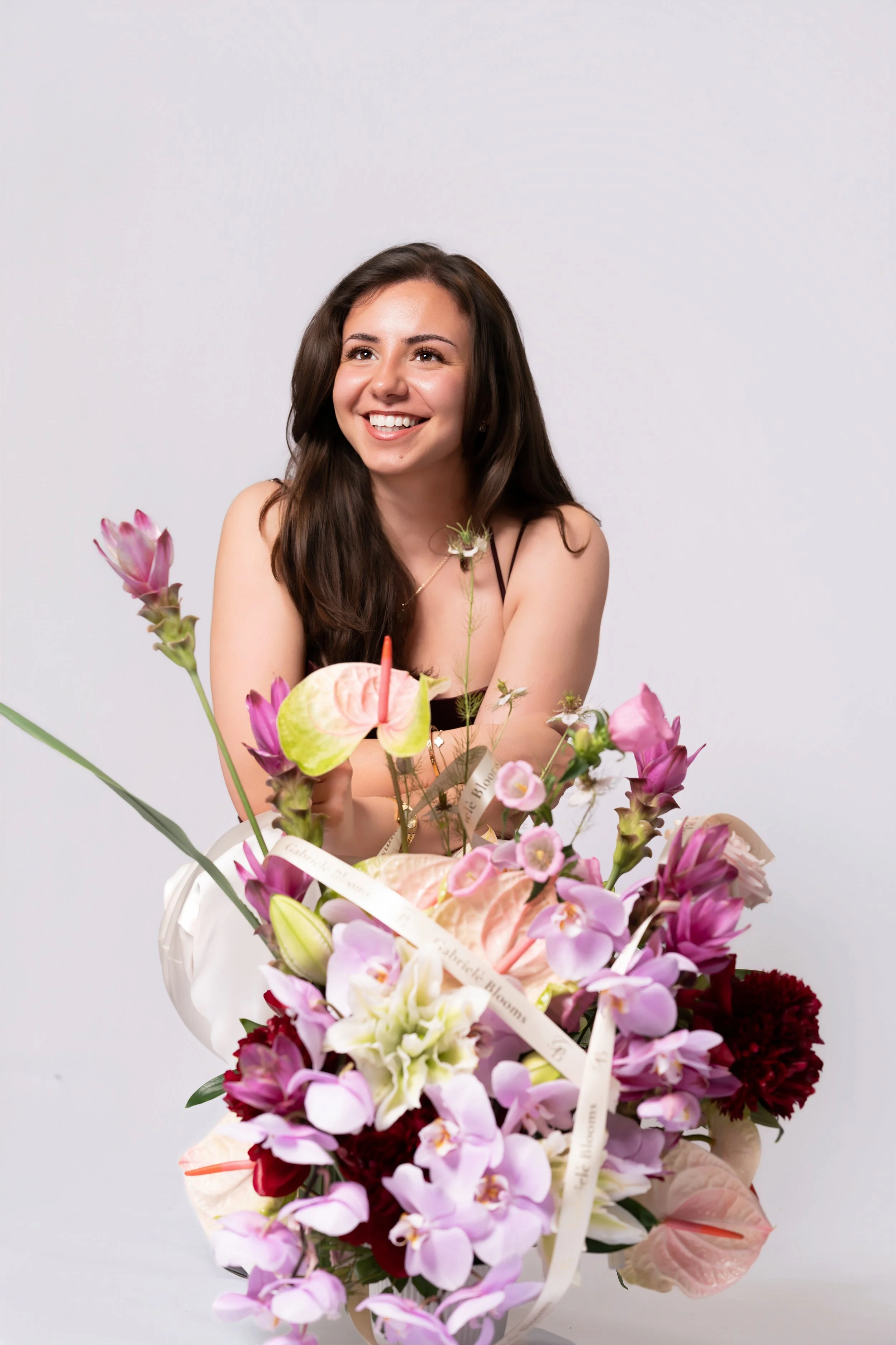 Gabriele Blooms smiling while sitting behind a large flower arrangement of pink, purple, white, and red flowers against a plain light background.