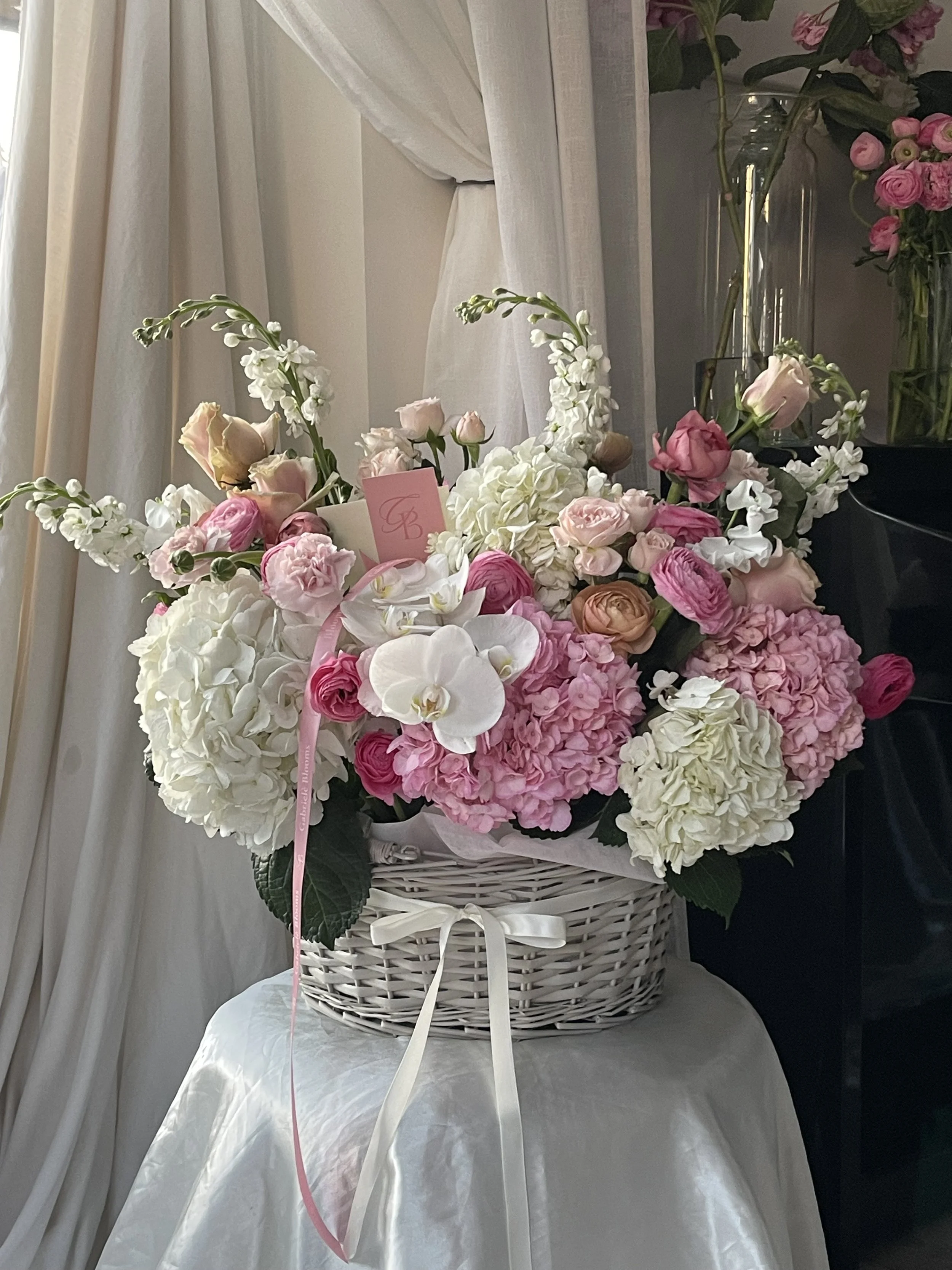 A white wicker basket filled with pink and white flowers, including roses and hydrangeas, with a pink ribbon tied around it, placed on a white surface with a sheer curtain background.