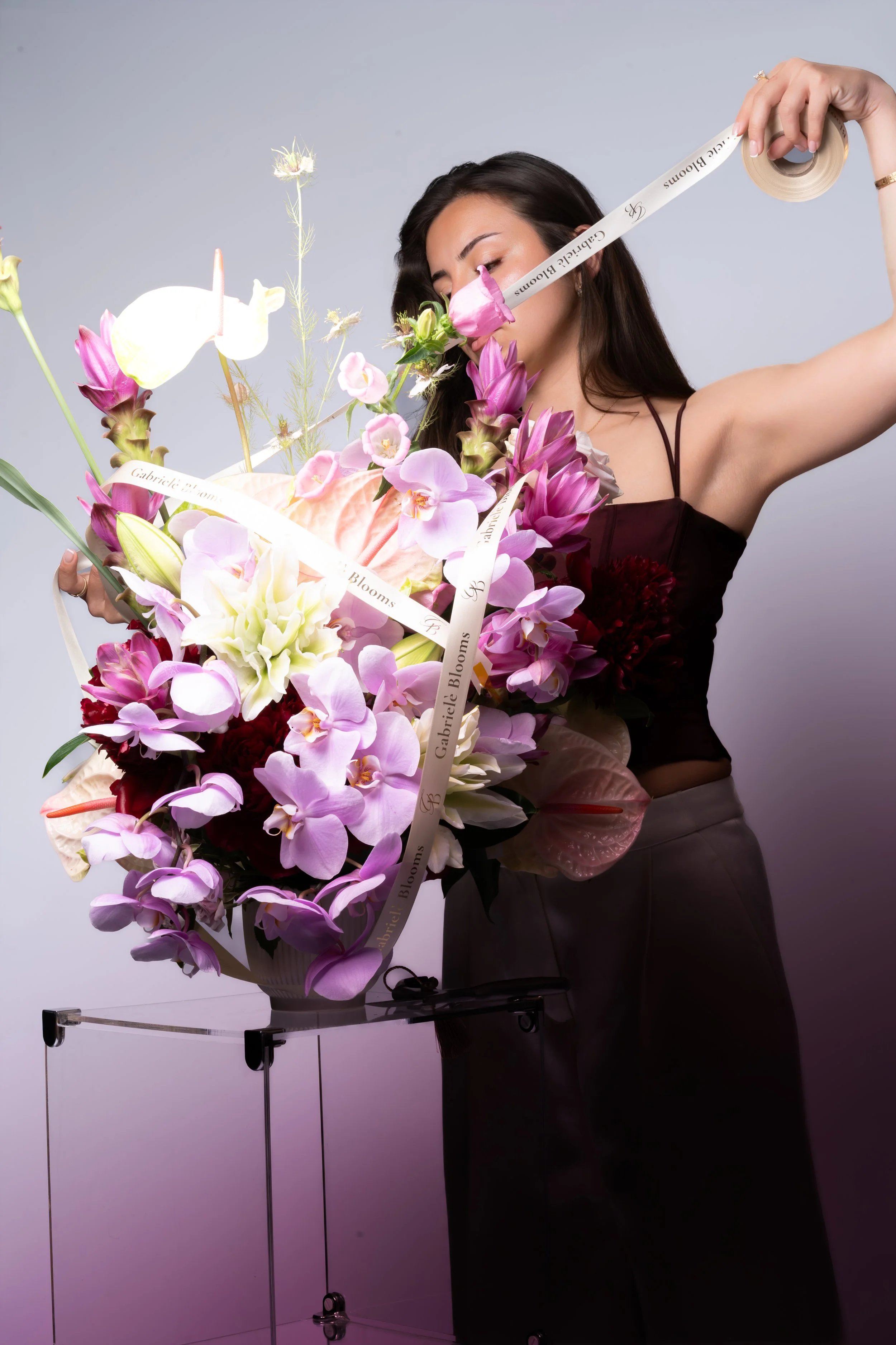 Gabriele Blooms photoshoot with a large arrangement of purple, pink, and white orchids, peonies ,anthurium, curcuma, lilies.Withribbons that have words like 'Gabriele Blooms' on them, standing in front of a light gray background.