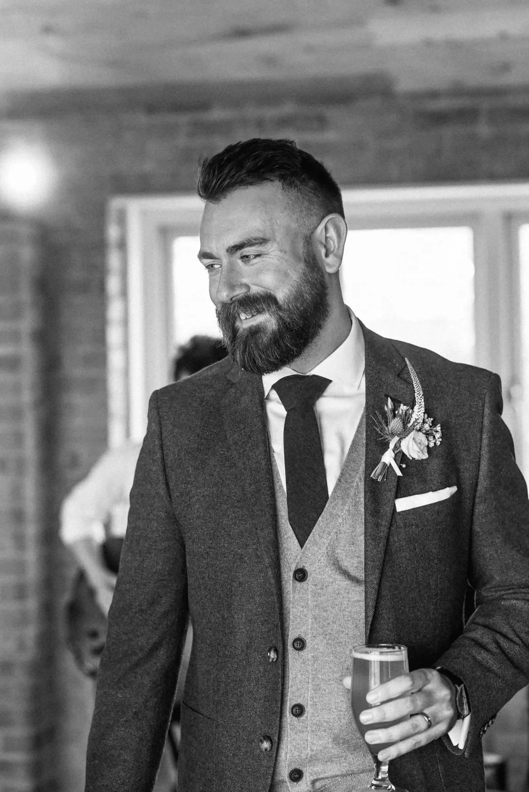 A man with a beard and styled hair, dressed in a suit with a boutonniere, holding a glass of wine, smiling at an indoor social event.