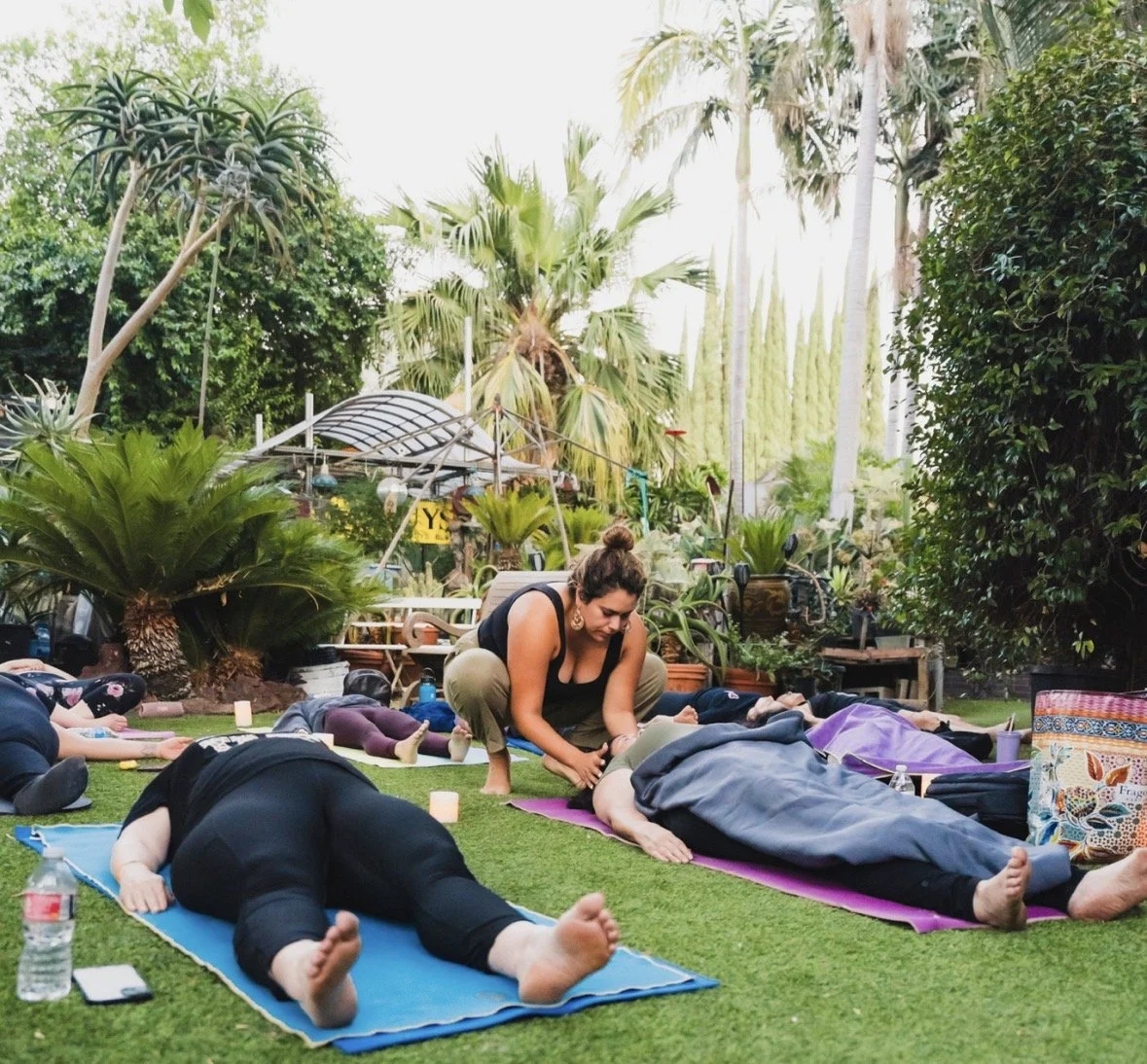 A group of people practicing yoga outdoors on mats in a lush garden with various plants and trees.