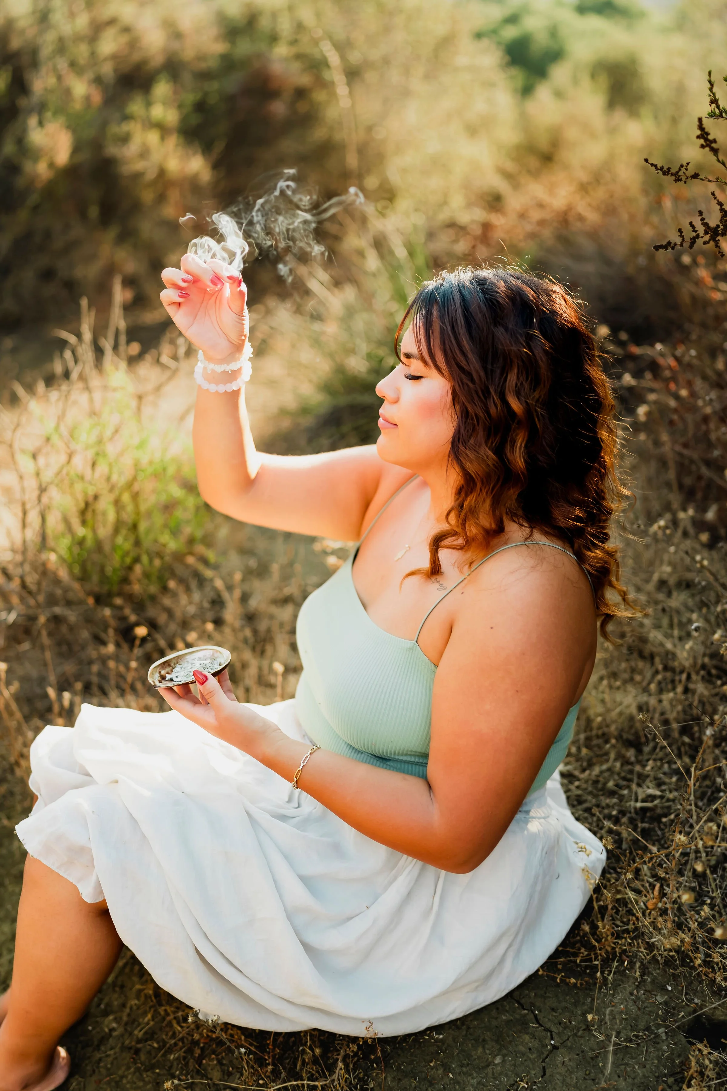 A woman sitting outdoors on a dirt patch with dry grass and bushes, holding an ice cream cone and enjoying a smoke, with her eyes closed and wearing a light blue tank top, white skirt, and jewelry, surrounded by warm sunlight.