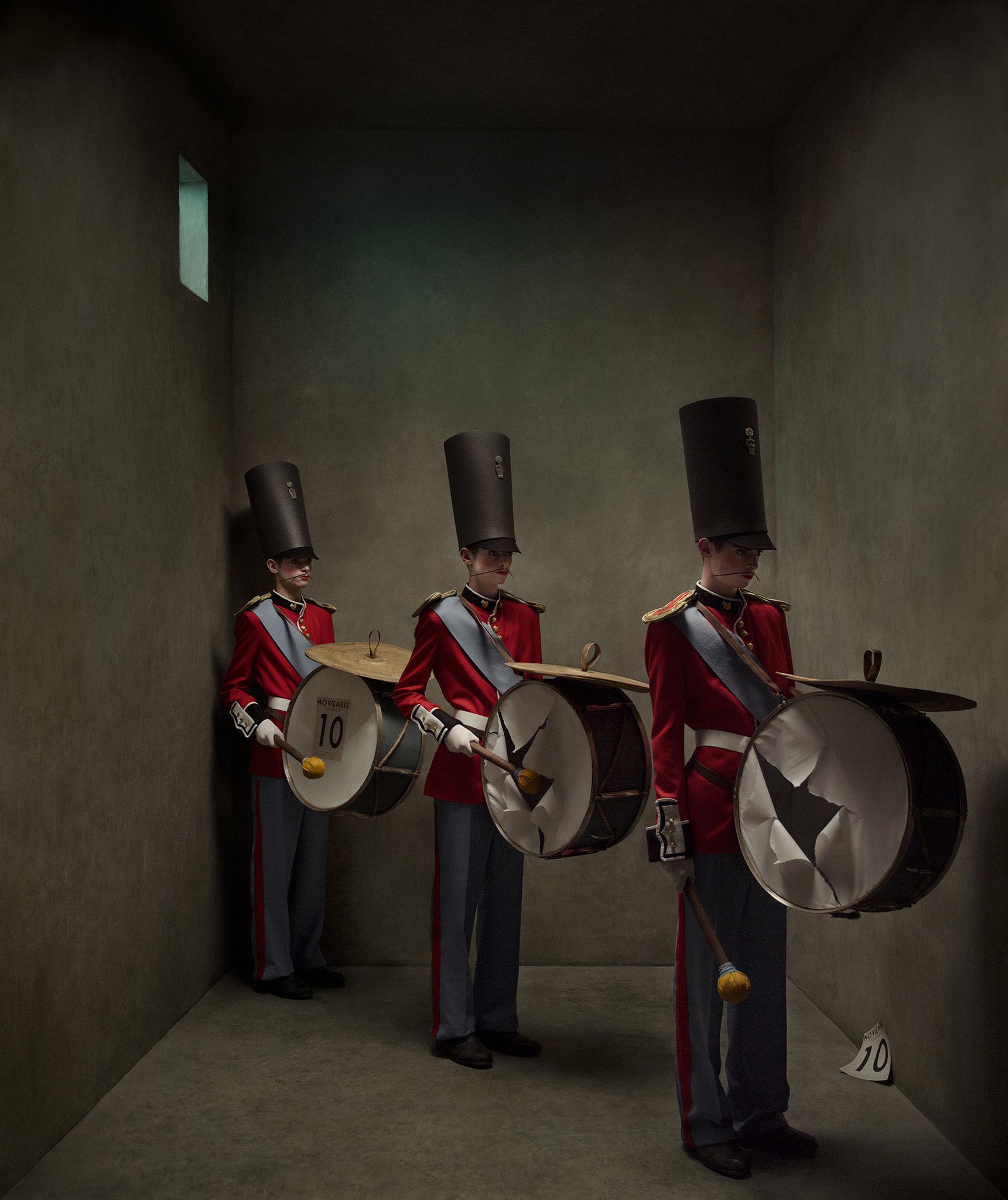 Four musicians dressed in red military-style uniforms with gray hats standing in dark, enclosed space, holding drums and drumsticks, with a small sign on the floor showing the number 10 and the word 'Noviembre'.