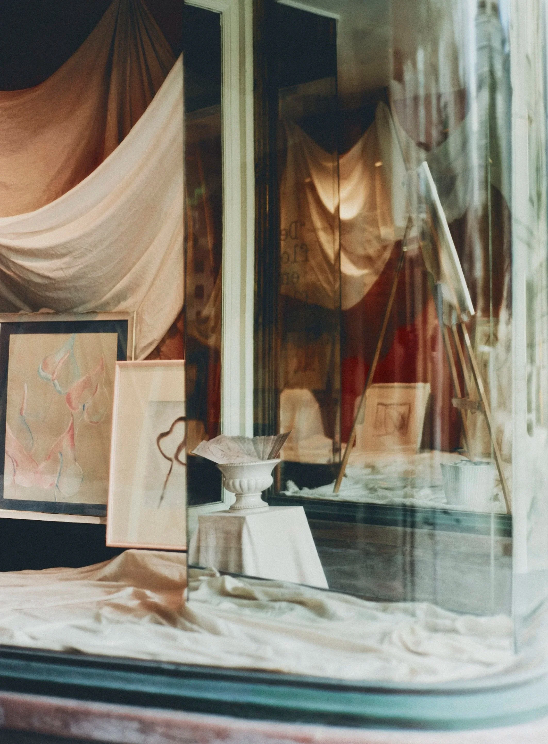 An art gallery window display with paintings and sculptures, including a pink and blue abstract artwork and a white classical urn with a drawing inside.