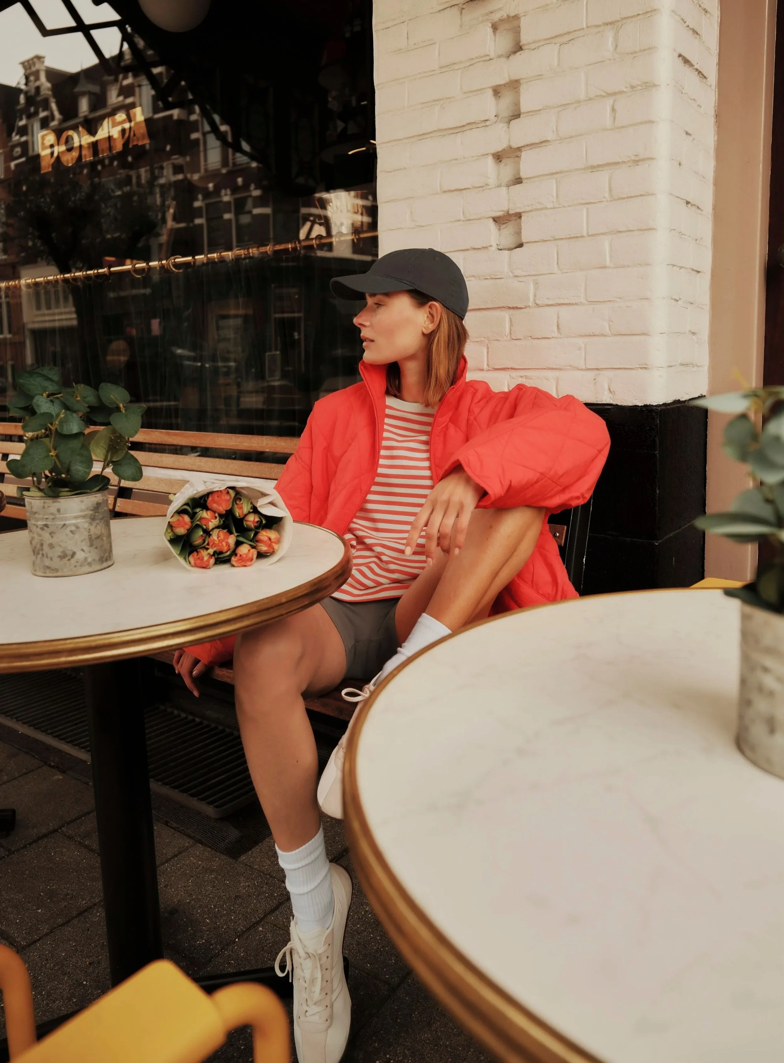 A woman with shoulder-length brown hair wearing a black cap, red jacket, and striped shirt is sitting outside at a cafe table. She is relaxing with her eyes closed. In front of her, there is a bouquet of orange roses and a small potted plant with gre