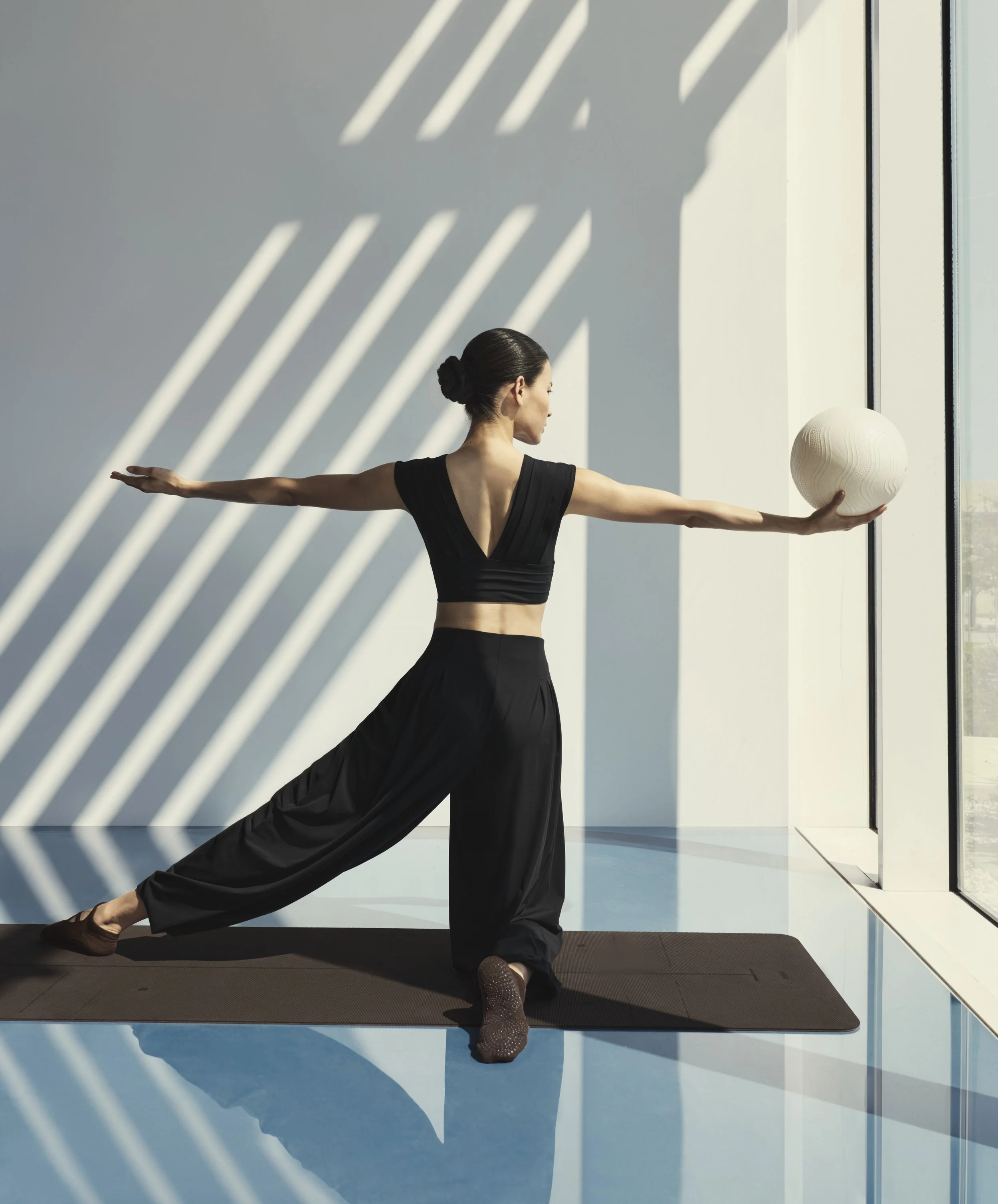 A woman in black workout clothing practices yoga or stretching exercises in front of a large window with sunlight casting shadows on the wall. She is holding a white textured ball in her right hand and is positioned on a yoga mat.