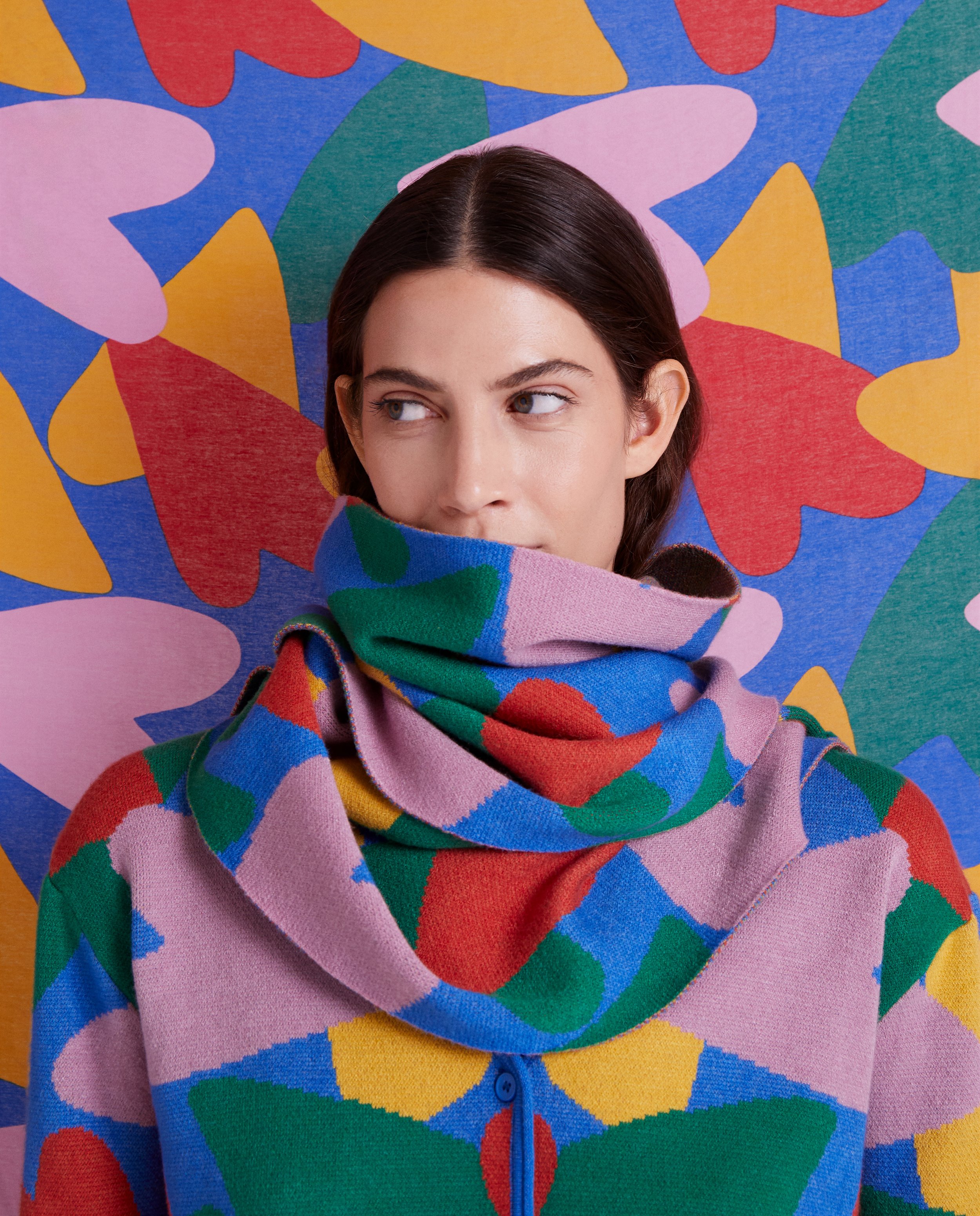 A woman with dark brown hair and blue eyes, wearing a colorful, abstract-patterned sweater with a high collar, standing in front of a vibrant, multicolored abstract mural background.